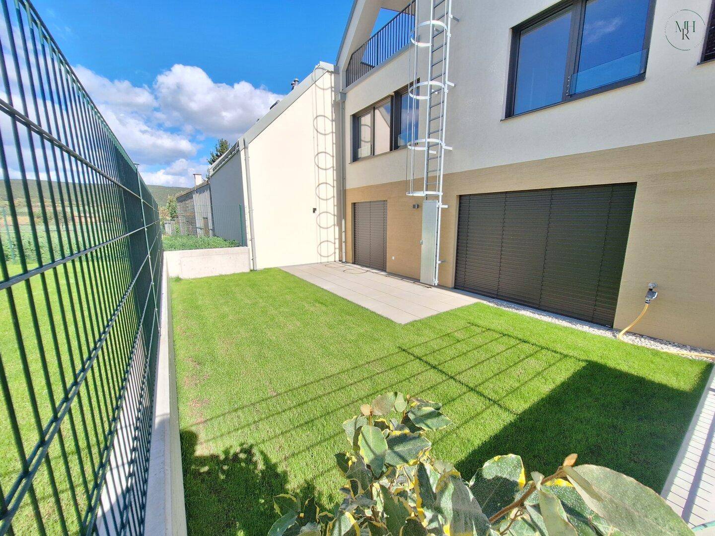 Sonniger Gartenbereich mit Rasen und kleiner Terrasse, umzäunt von einem Gitterzaun.
