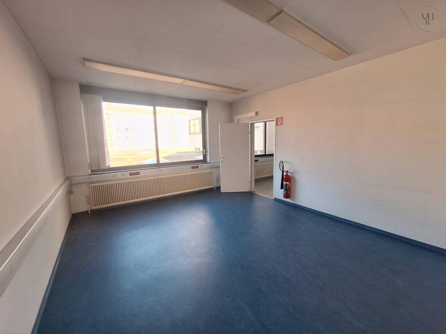 Leerer Büroraum mit blauer Bodenfläche, großem Fenster und einem Feuerlöscher an der Wand. Die Beleuchtung ist funktional.