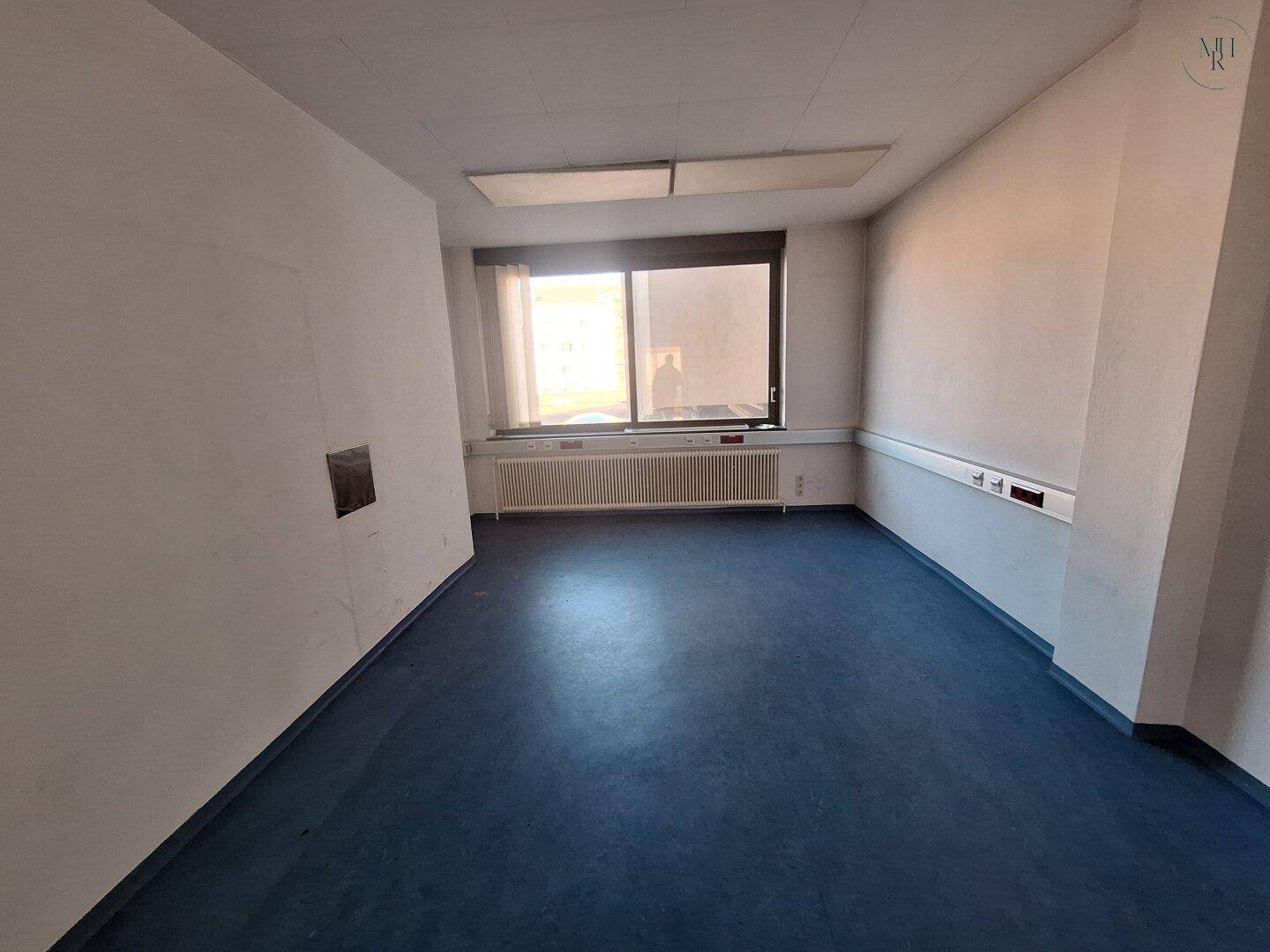 Schlichter Büroraum mit blauem Boden, einem Fenster mit Jalousien und einem Heizkörper unter der Fensterbank.