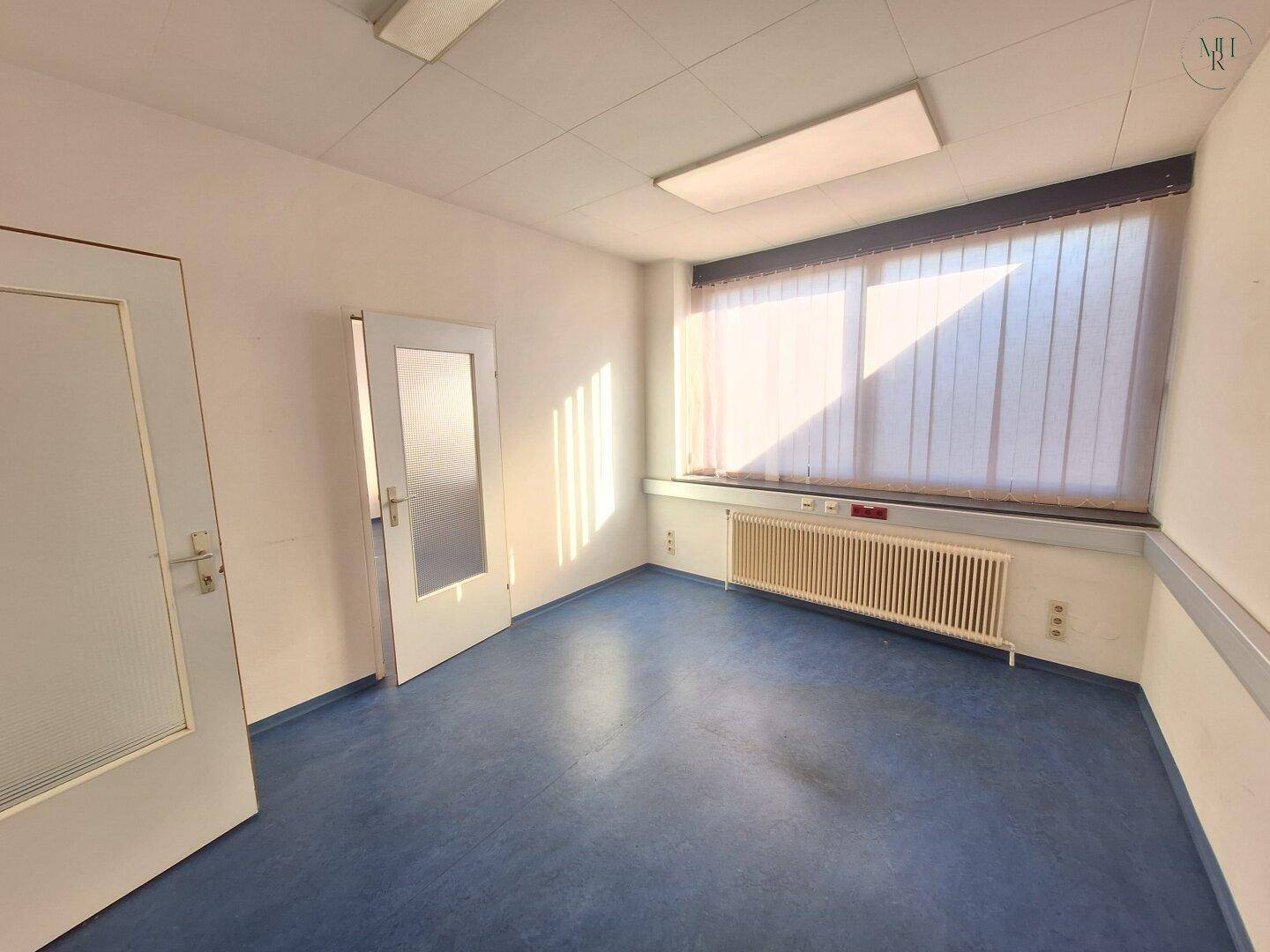 Freundlicher Büroraum mit blauer Bodenfläche, zwei Türen und einem Fenster mit vertikalen Jalousien. Viel Sonnenlicht fällt herein.
