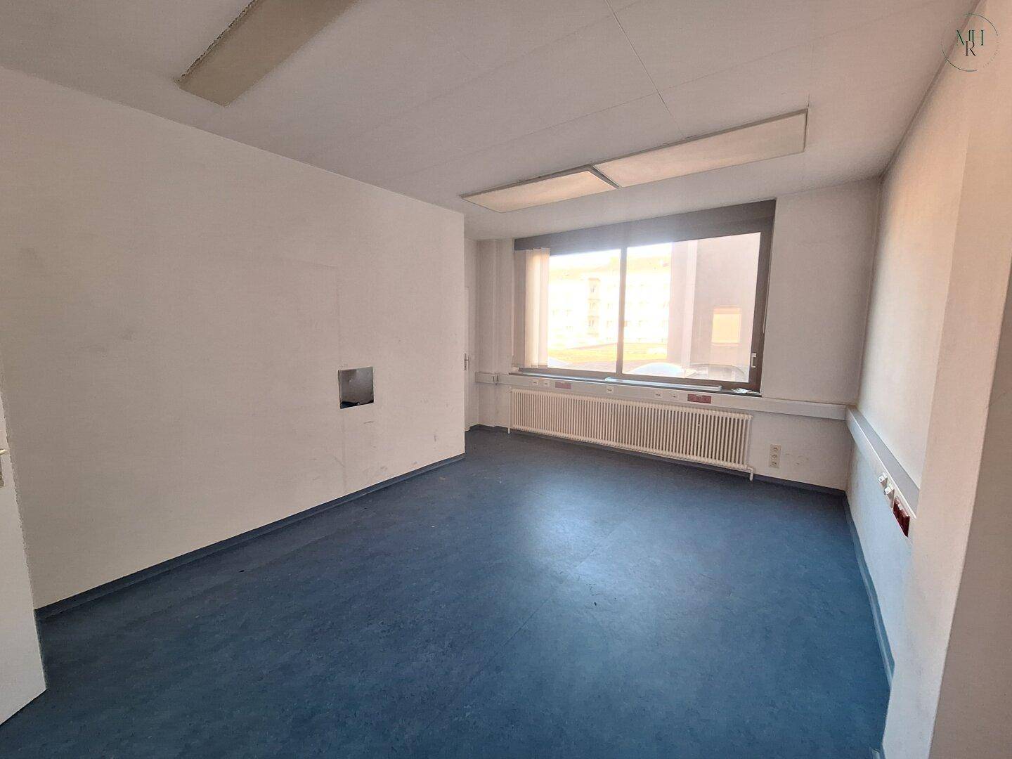 Heller Büroraum mit blauer Bodenfläche, großem Fenster und einem Heizkörper unter der Fensterbank. Die Wände sind weiß.