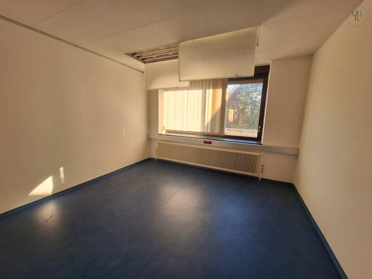 Einfacher Büroraum mit blauem Boden, einem Fenster mit Jalousien und einem Heizkörper. Die Deckenplatten sind sichtbar.