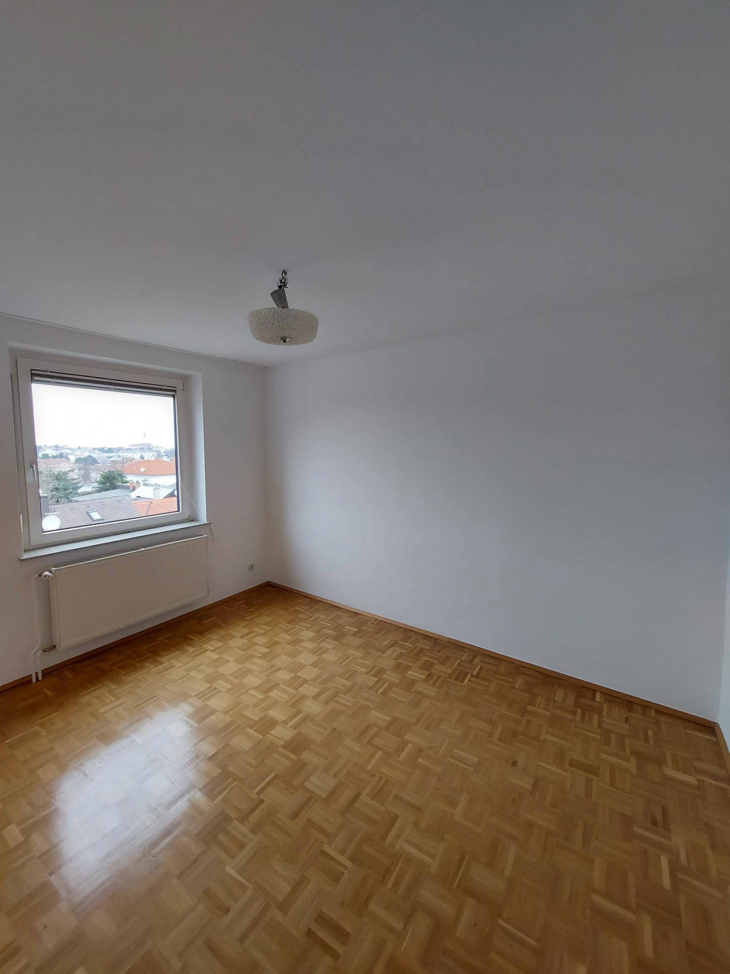 Leeres Schlafzimmer mit Parkettboden, weißen Wänden und einem Fenster mit Heizkörper darunter.