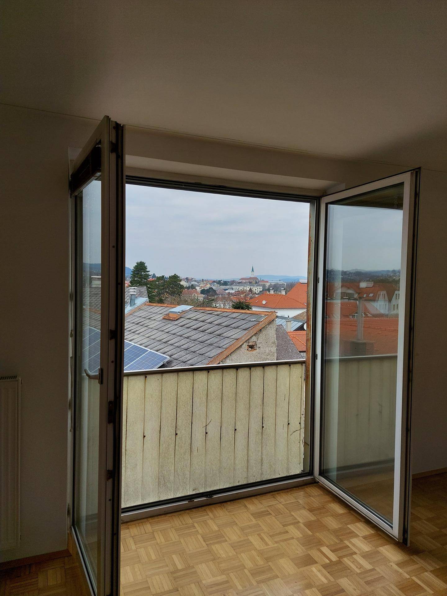 Offenes Fenster in einem hellen Raum mit Parkettboden und Blick über die Dächer der Stadt.