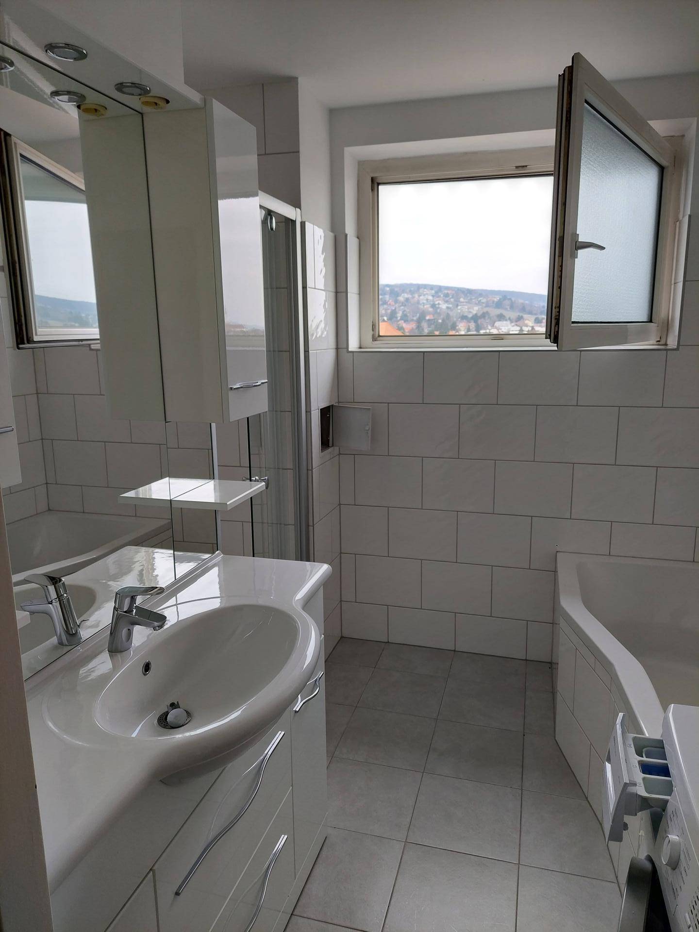 Helles Badezimmer mit weißen Fliesen, Waschbecken, Spiegelschrank und Badewanne am Fenster.