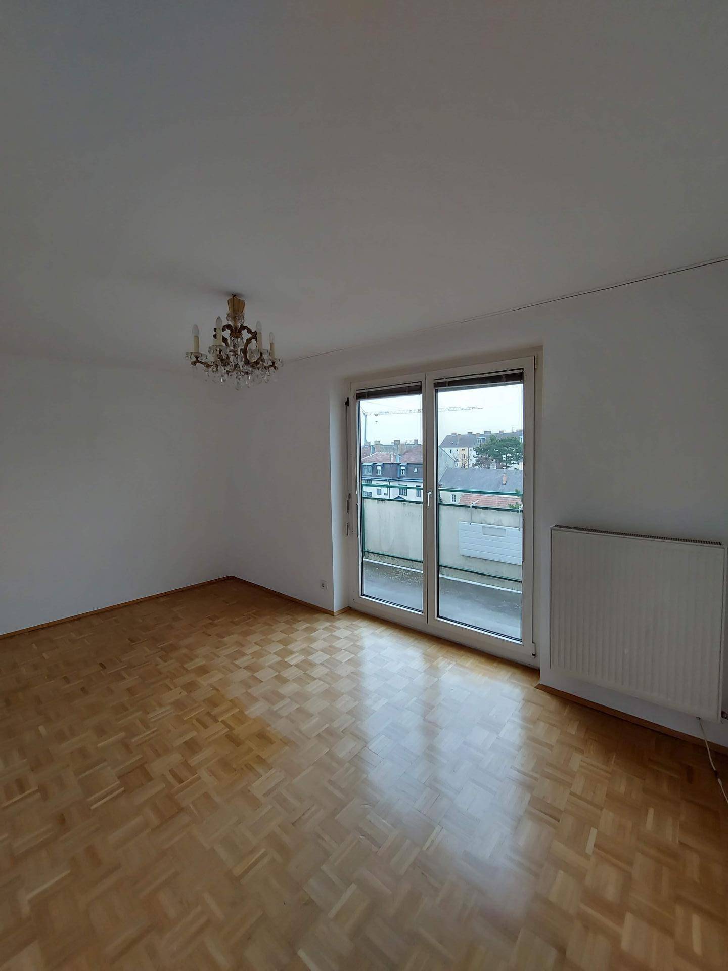 Helles Wohnzimmer mit Parkettboden, weißen Wänden und großem Fenster mit Zugang zu einem Balkon.