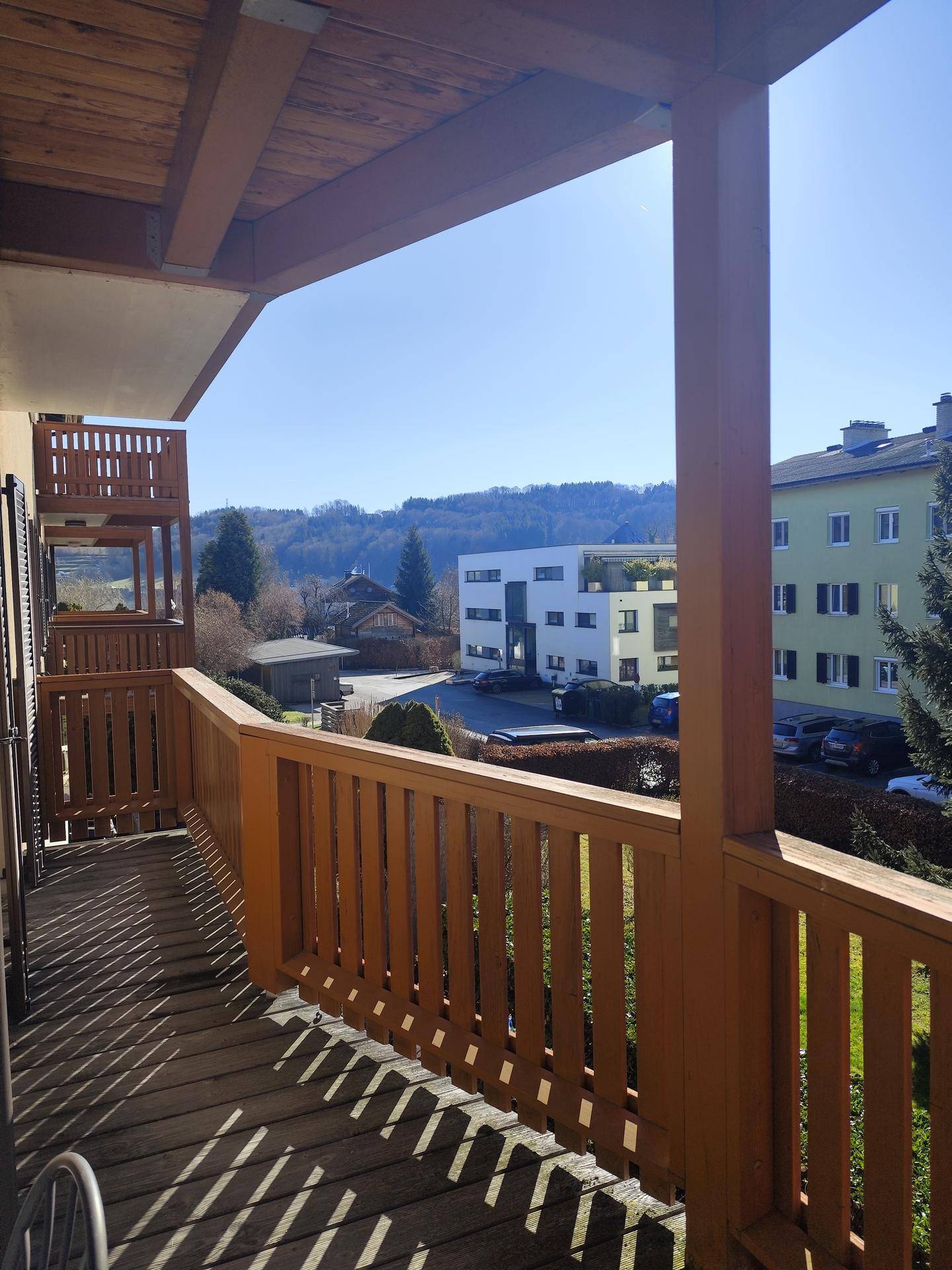 Sonniger Balkon mit Holzgeländer und Blick auf die umliegende Wohngegend und grüne Hügel.