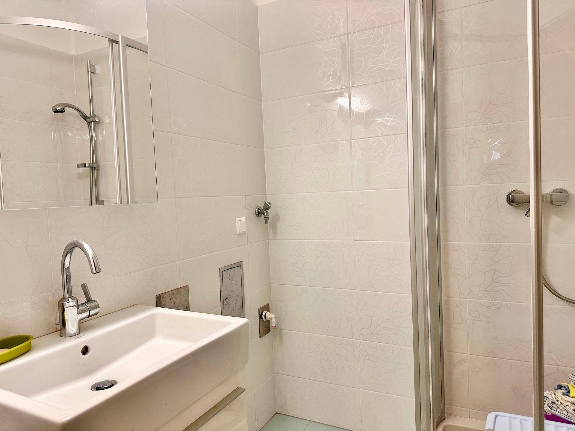 Badezimmer mit Waschbecken, Spiegel und einer Duschkabine, hell gefliest.