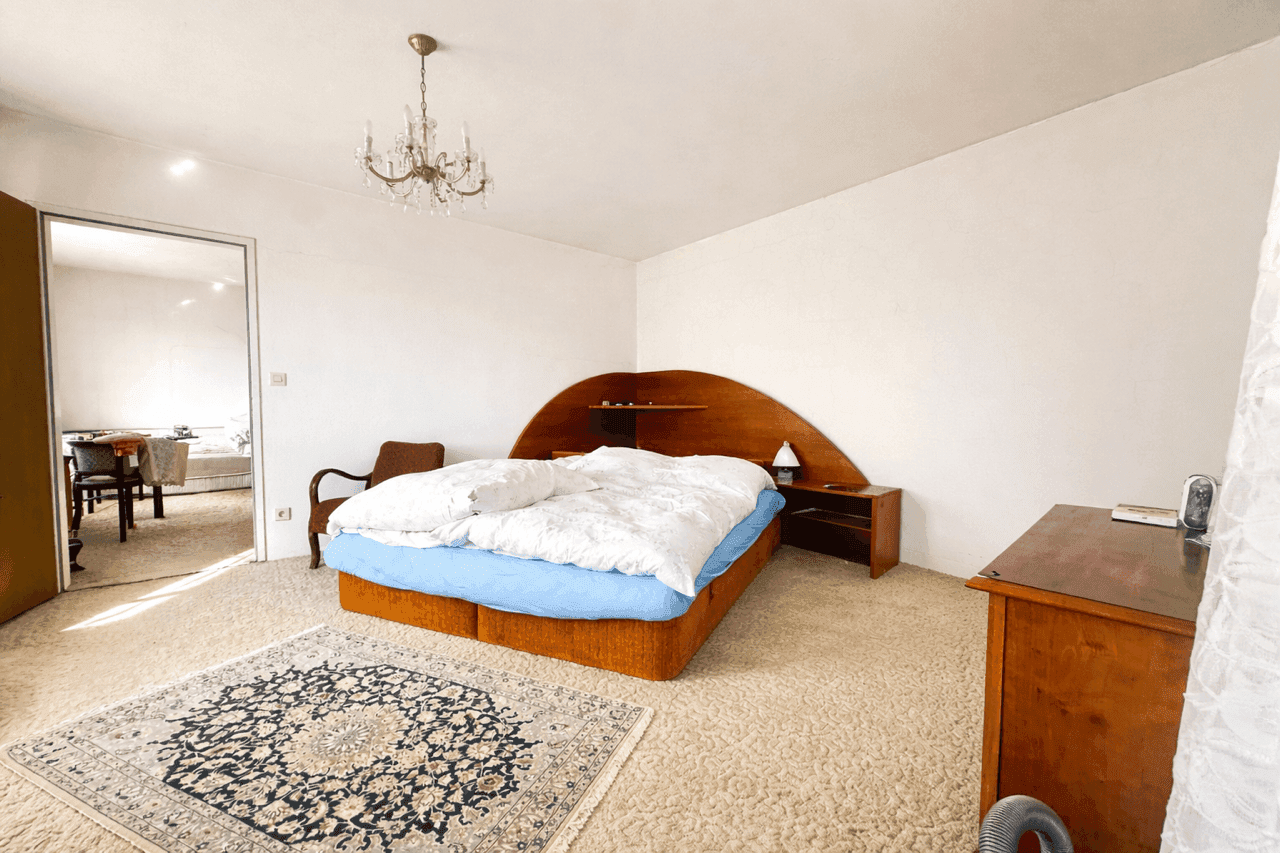 Geräumiges Schlafzimmer mit Teppichboden, großem Bett und Kronleuchter, angrenzend an einen weiteren Raum.