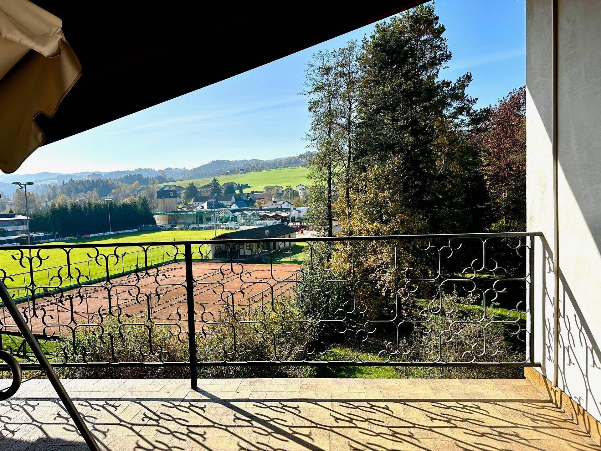 Sonniger Balkon mit Blick auf die umliegende Natur, einen Tennisplatz und ferne Hügel.