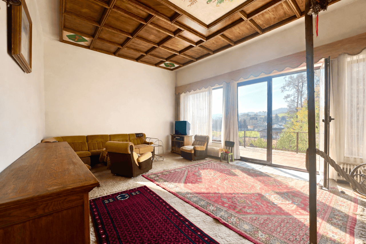 Geräumiges Wohnzimmer mit Holzkassettendecke, Teppichboden und Zugang zu einer großen Terrasse.