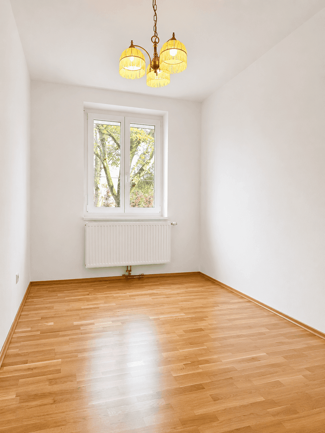 Leerer Raum mit Parkettboden, weißen Wänden und einem Fenster, das viel Tageslicht hereinlässt.
