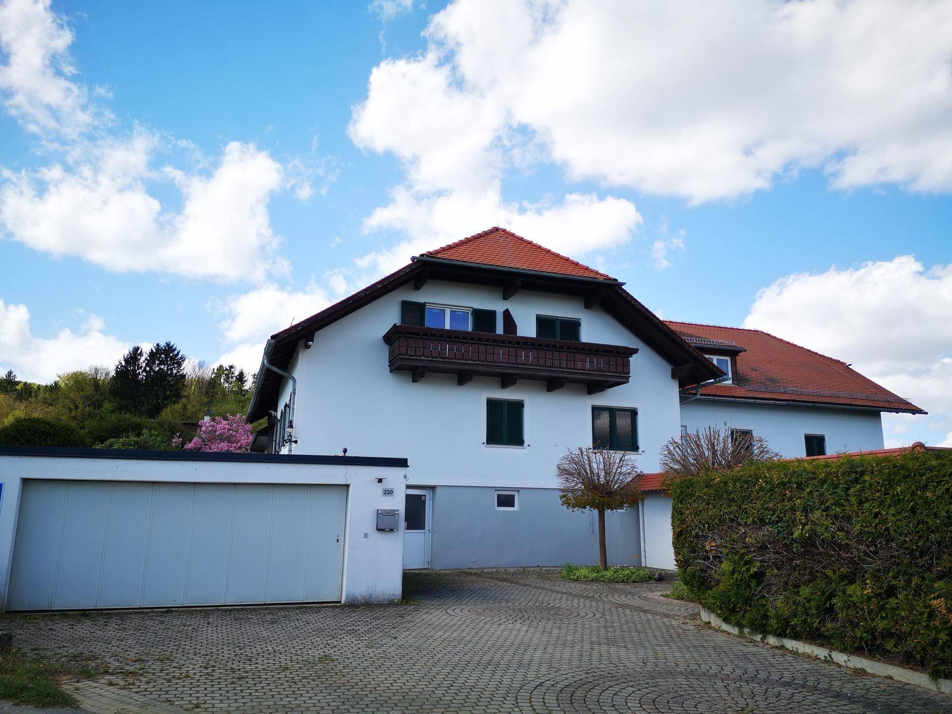 Wohnhaus mit Garage und gepflasterter Auffahrt, umgeben von Hecken und Bäumen unter blauem Himmel.