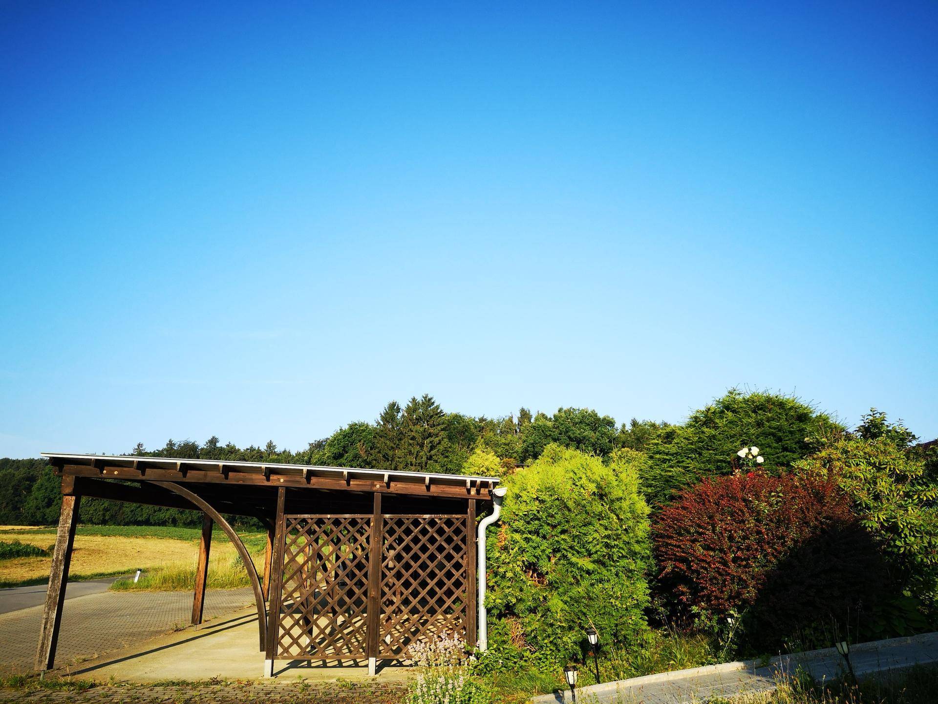 Holzcarport mit Gitterelementen, umgeben von üppiger Vegetation und einem Feld unter klarem Himmel.