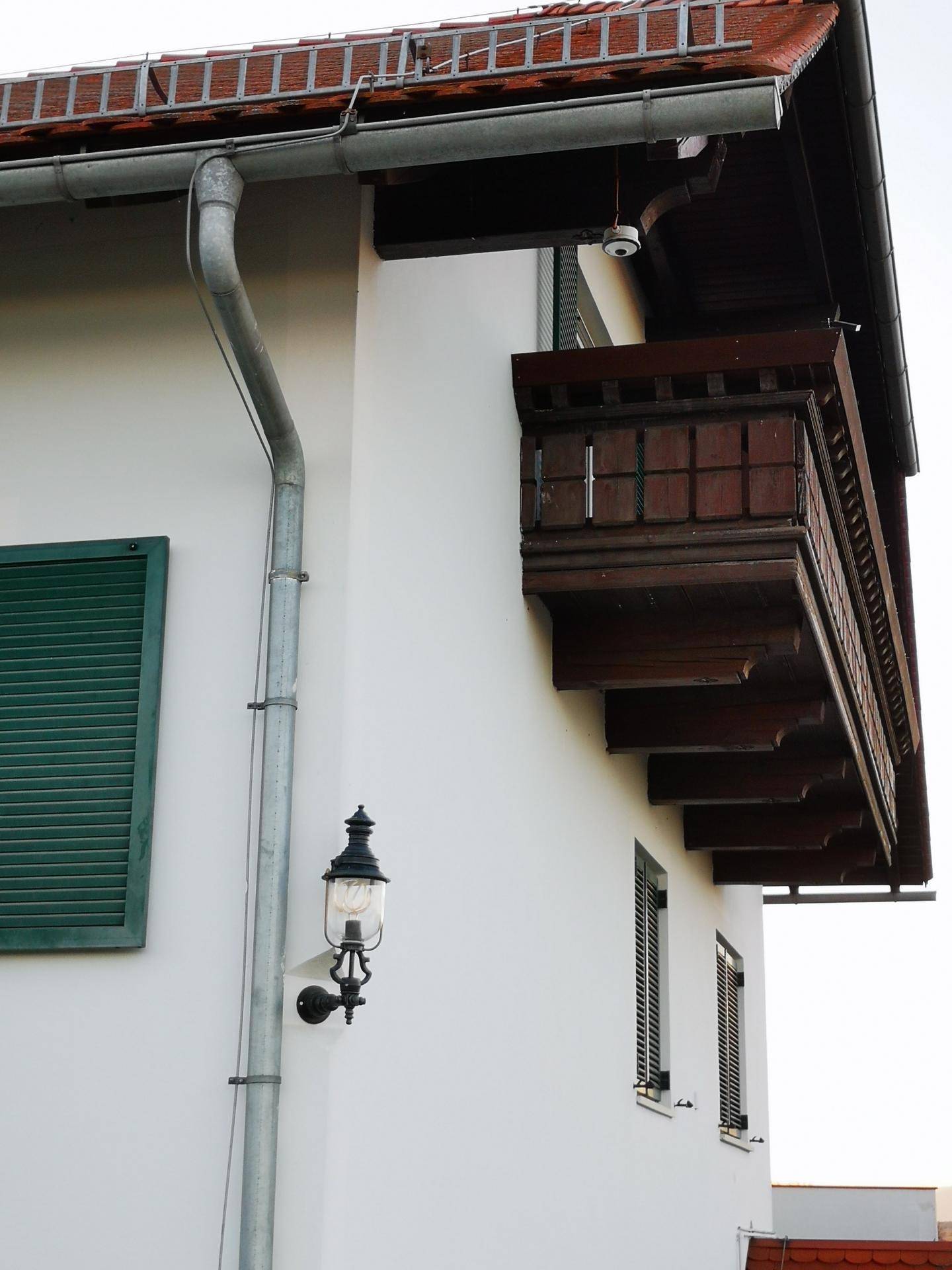 Detailansicht einer Hausfassade mit Holzbalkon, Regenrinne und einer Außenleuchte.