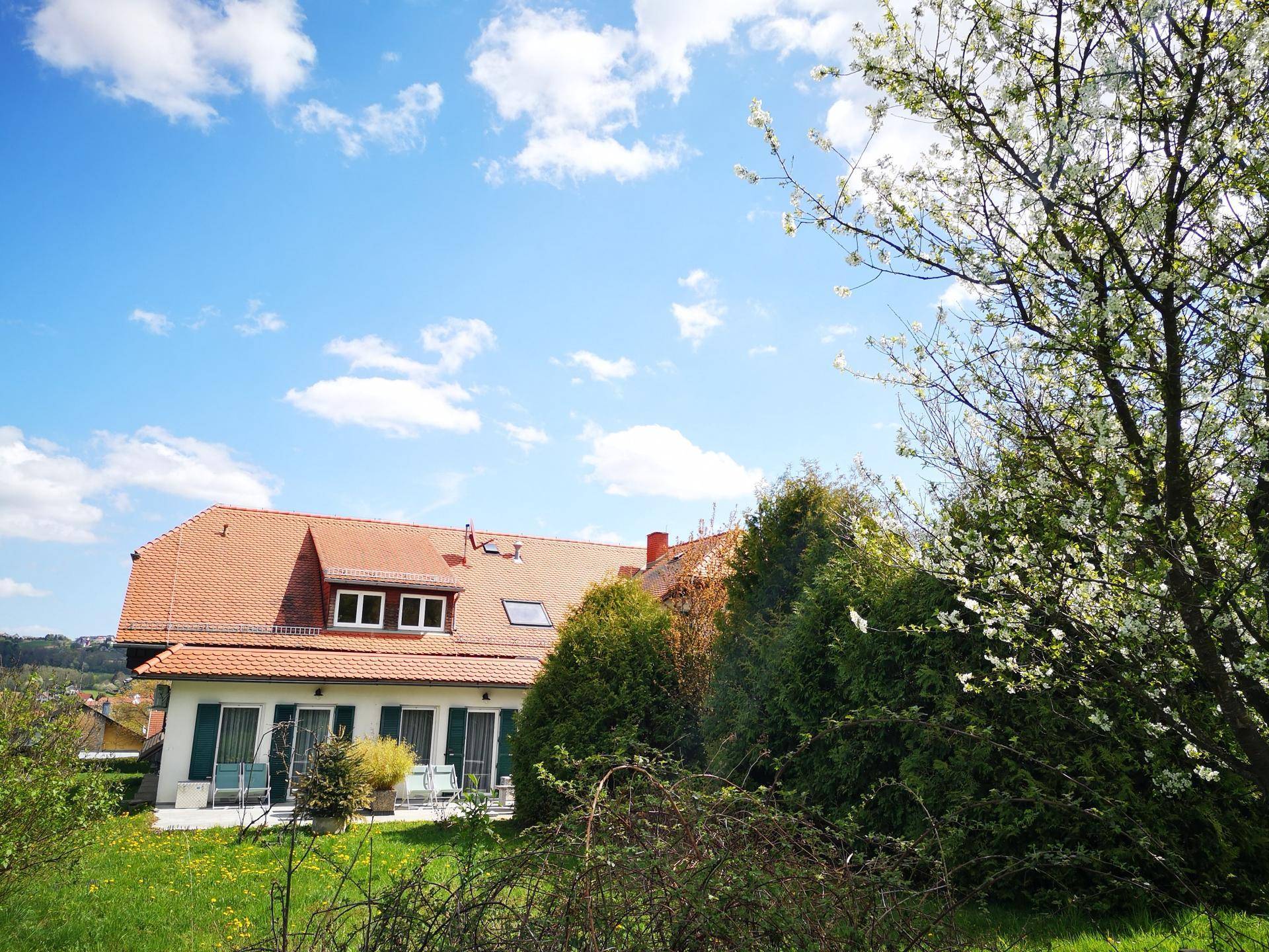Großes Haus mit rotem Ziegeldach und grünen Fensterläden, umgeben von einem weitläufigen Garten und Bäumen.