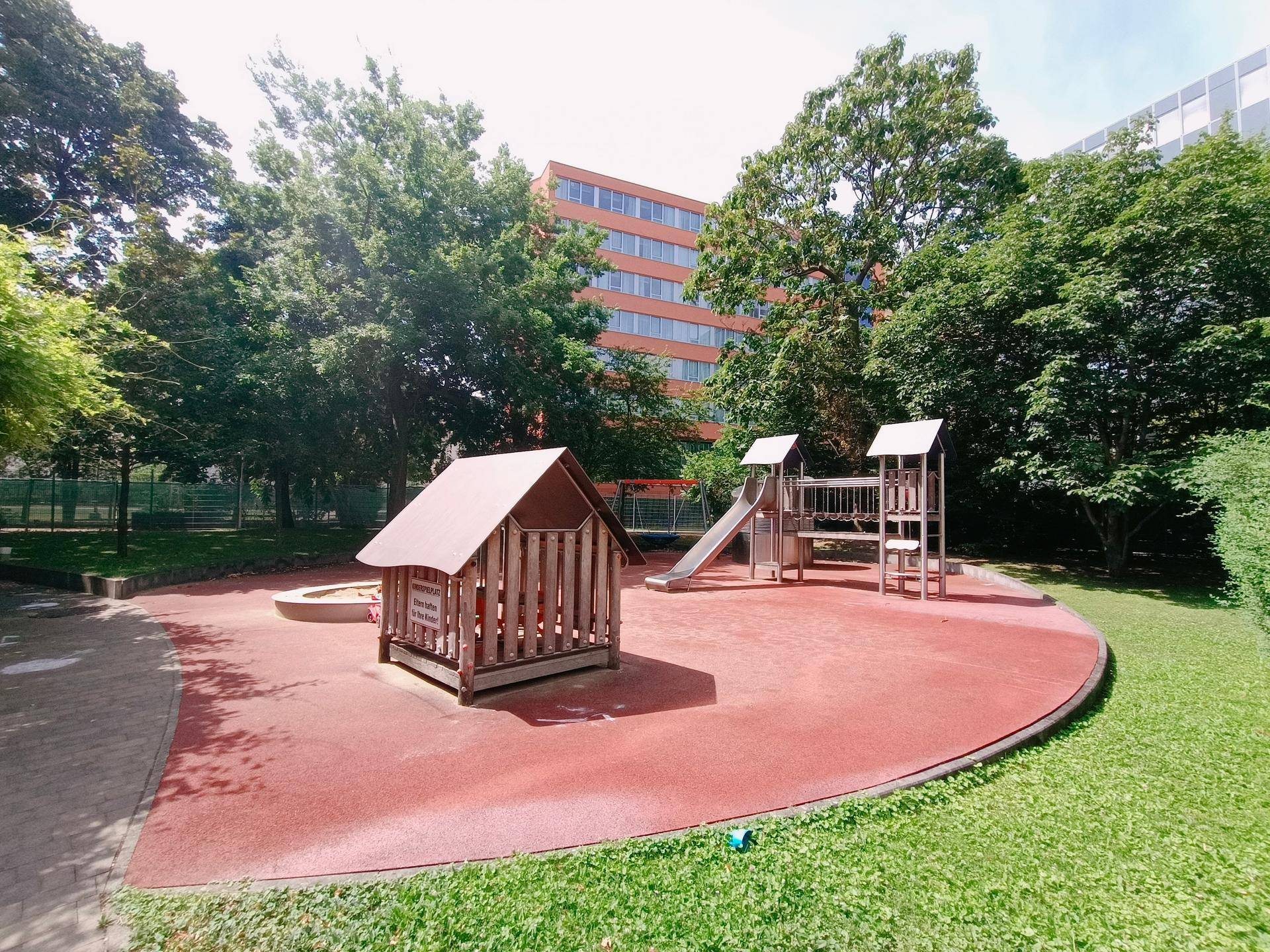Kinderspielplatz mit Rutsche, Klettergerüst und Spielhaus, umgeben von viel Grün.