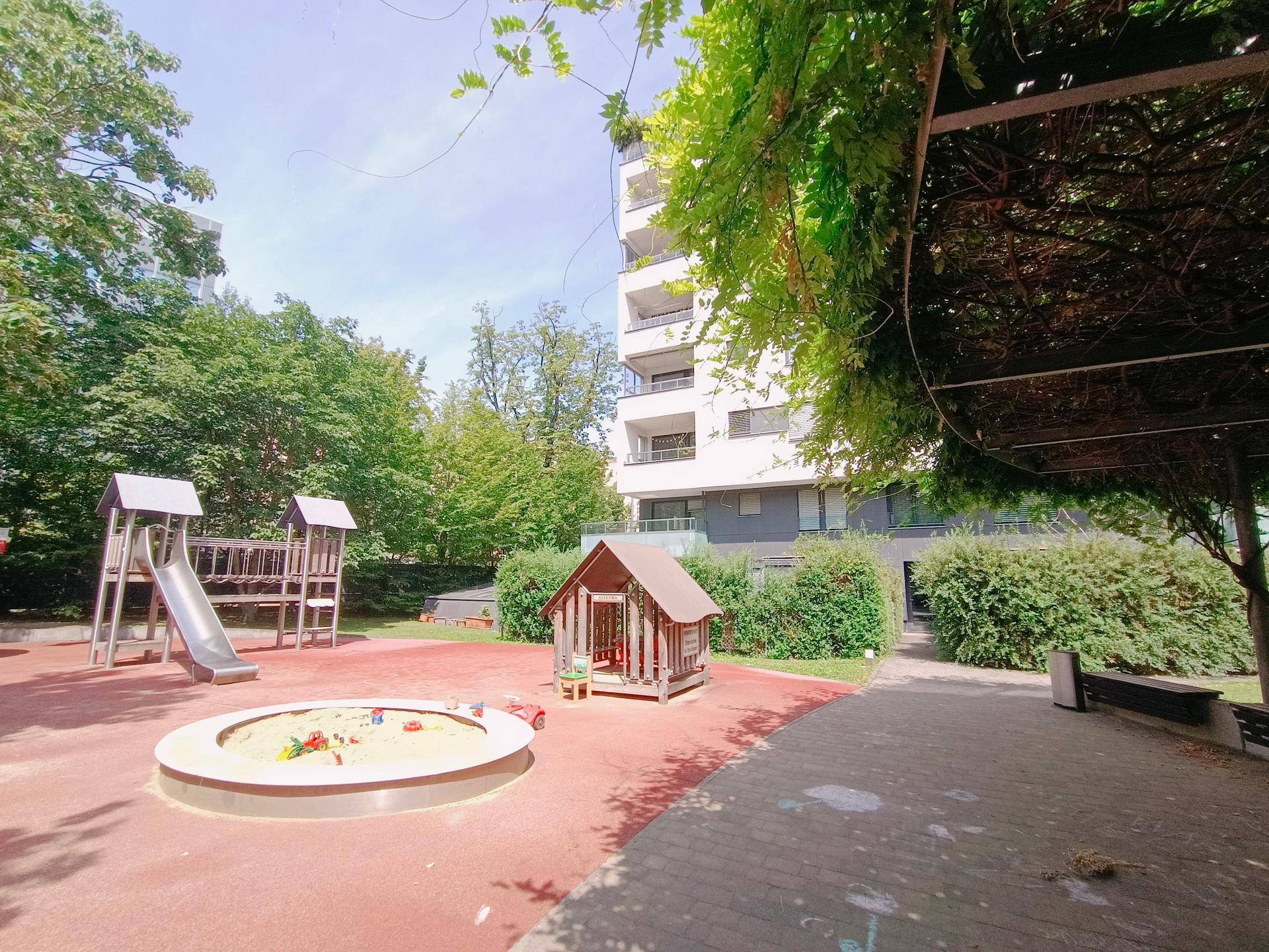 Spielplatz mit Sandkasten und Spielgeräten, umgeben von Bäumen und einem modernen Wohngebäude.