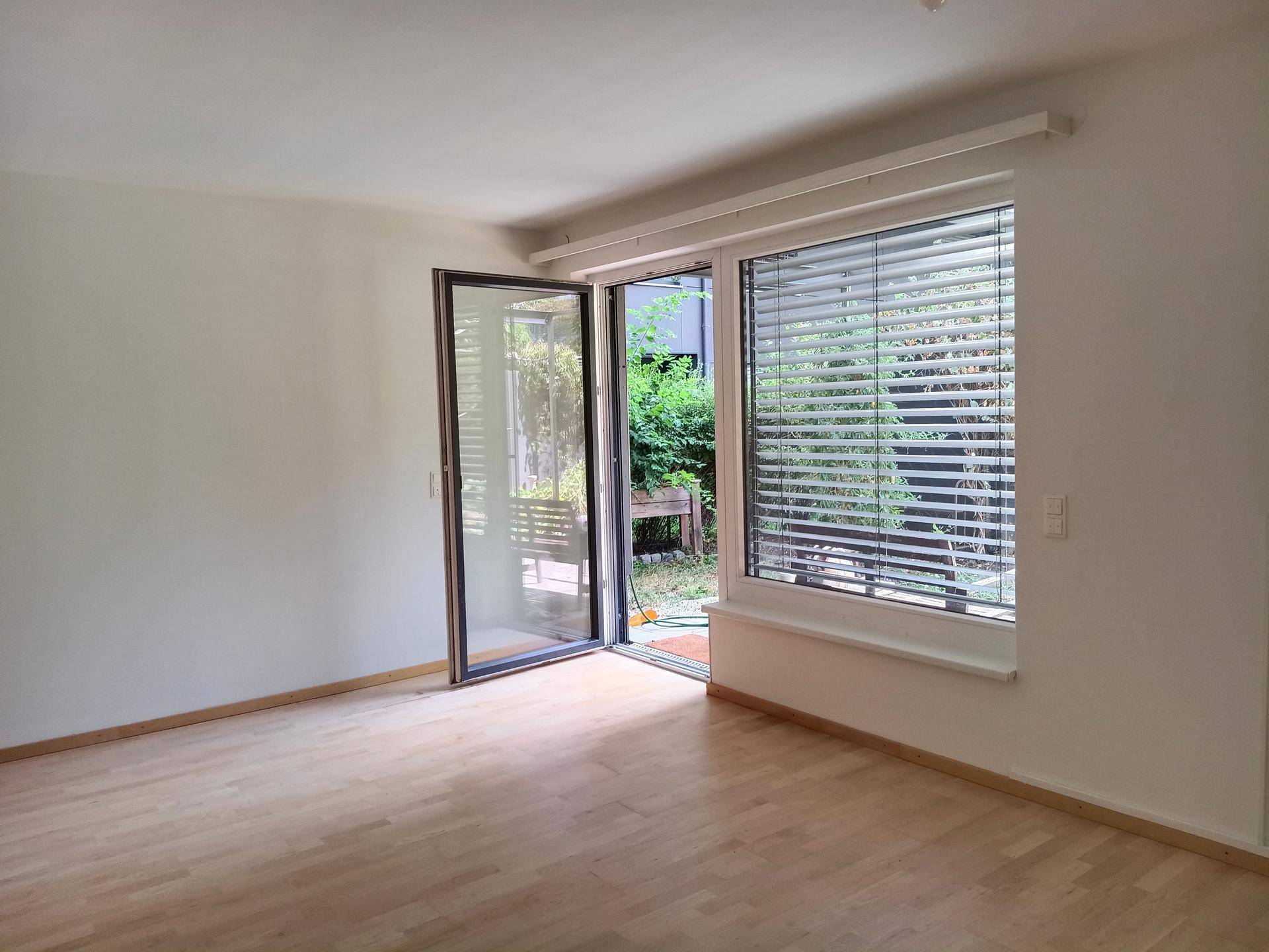 Heller Wohnraum mit Holzboden und Zugang zu einem Außenbereich, durch eine weit geöffnete Glastür.
