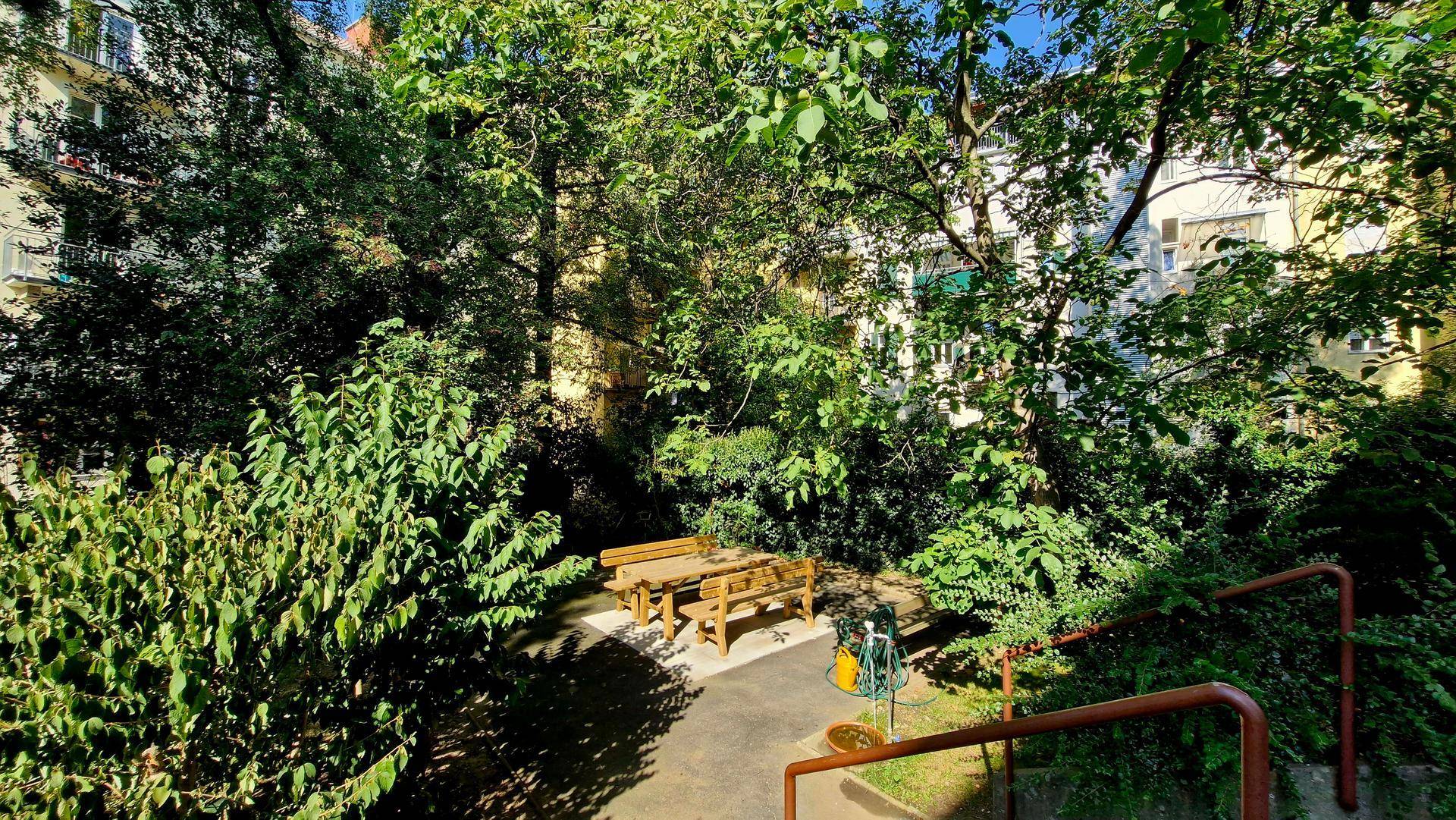 Grüner Gemeinschaftsgarten mit Holztisch und Bänken, umgeben von Bäumen und Sträuchern an einem sonnigen Tag.