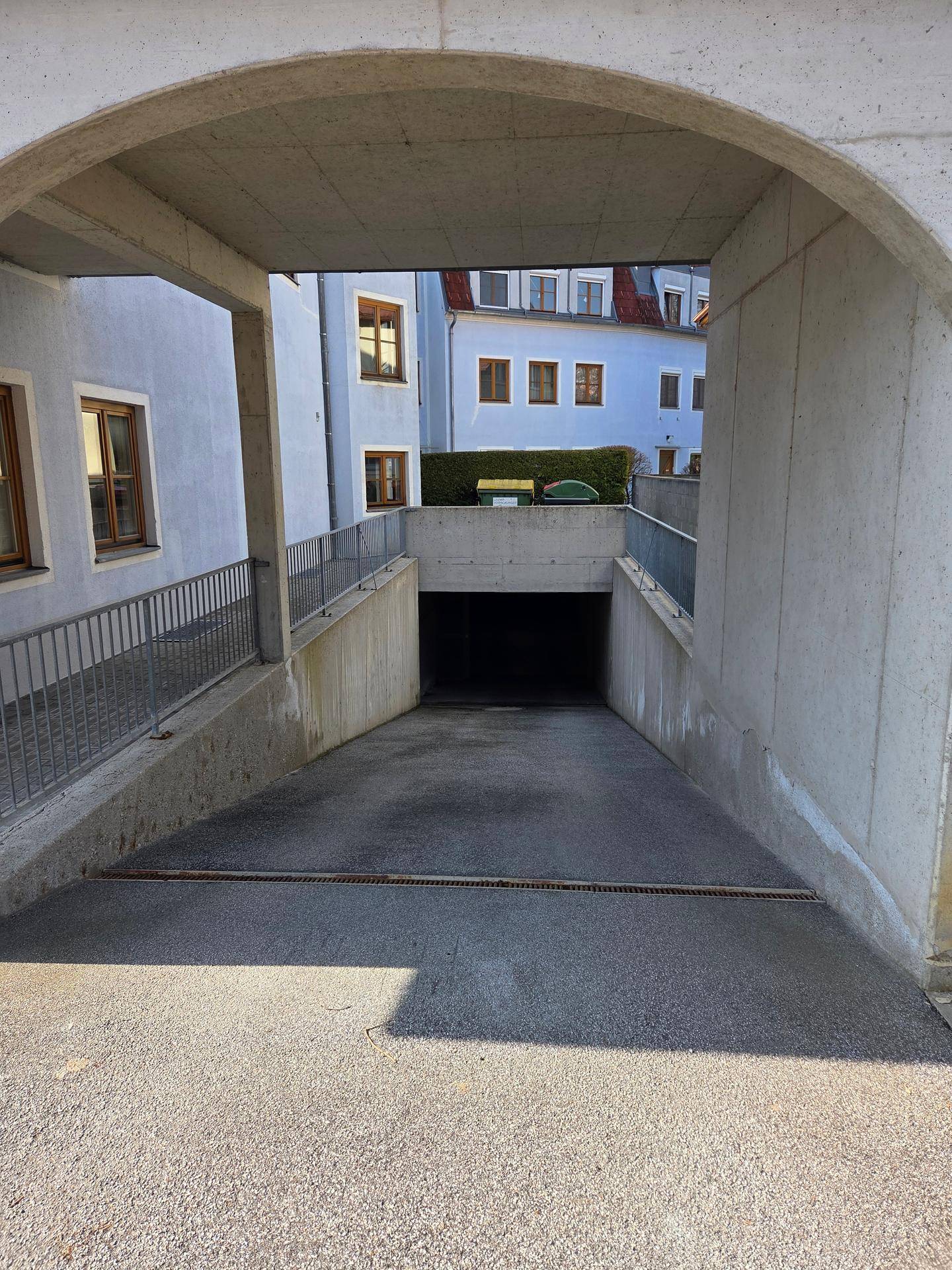 Zufahrt zur Tiefgarage unter einem Bogen mit umliegenden Gebäuden an einem sonnigen Tag.