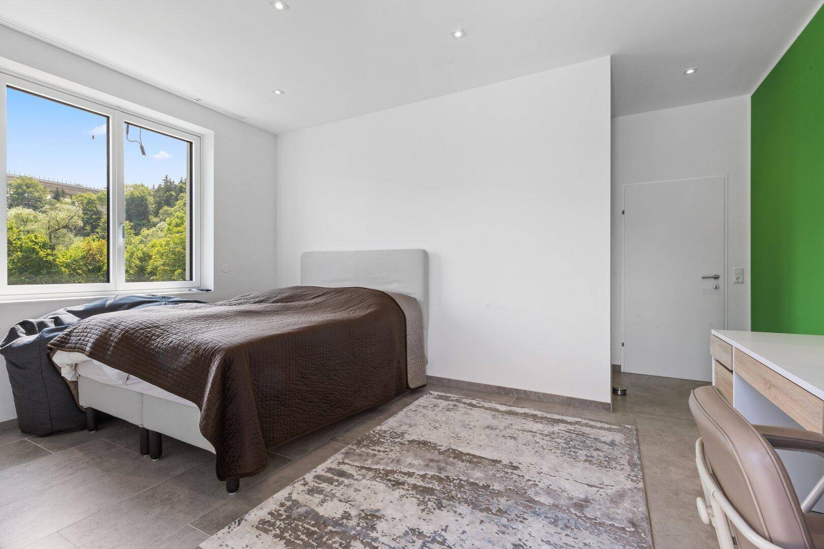 Schlafzimmer mit Bett, Schreibtisch und einem Fenster, das viel Tageslicht hereinlässt.
