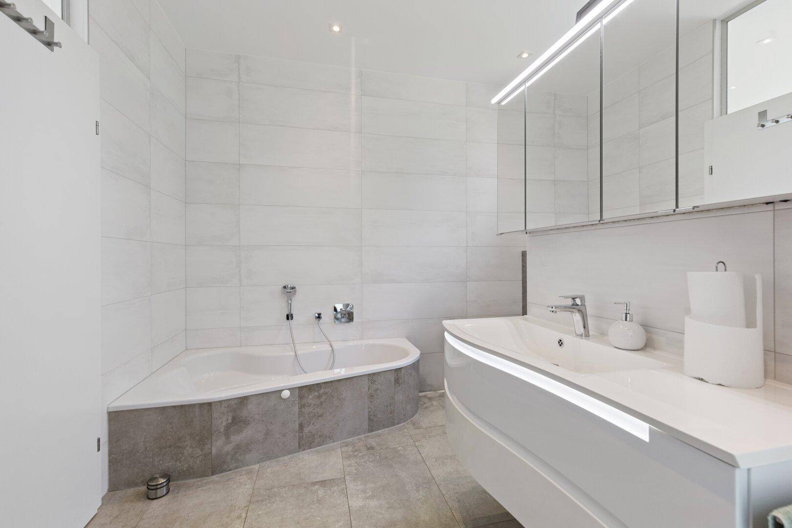Modernes Badezimmer mit Eckbadewanne, elegantem Waschtisch und hellen Fliesen an Wänden und Boden.