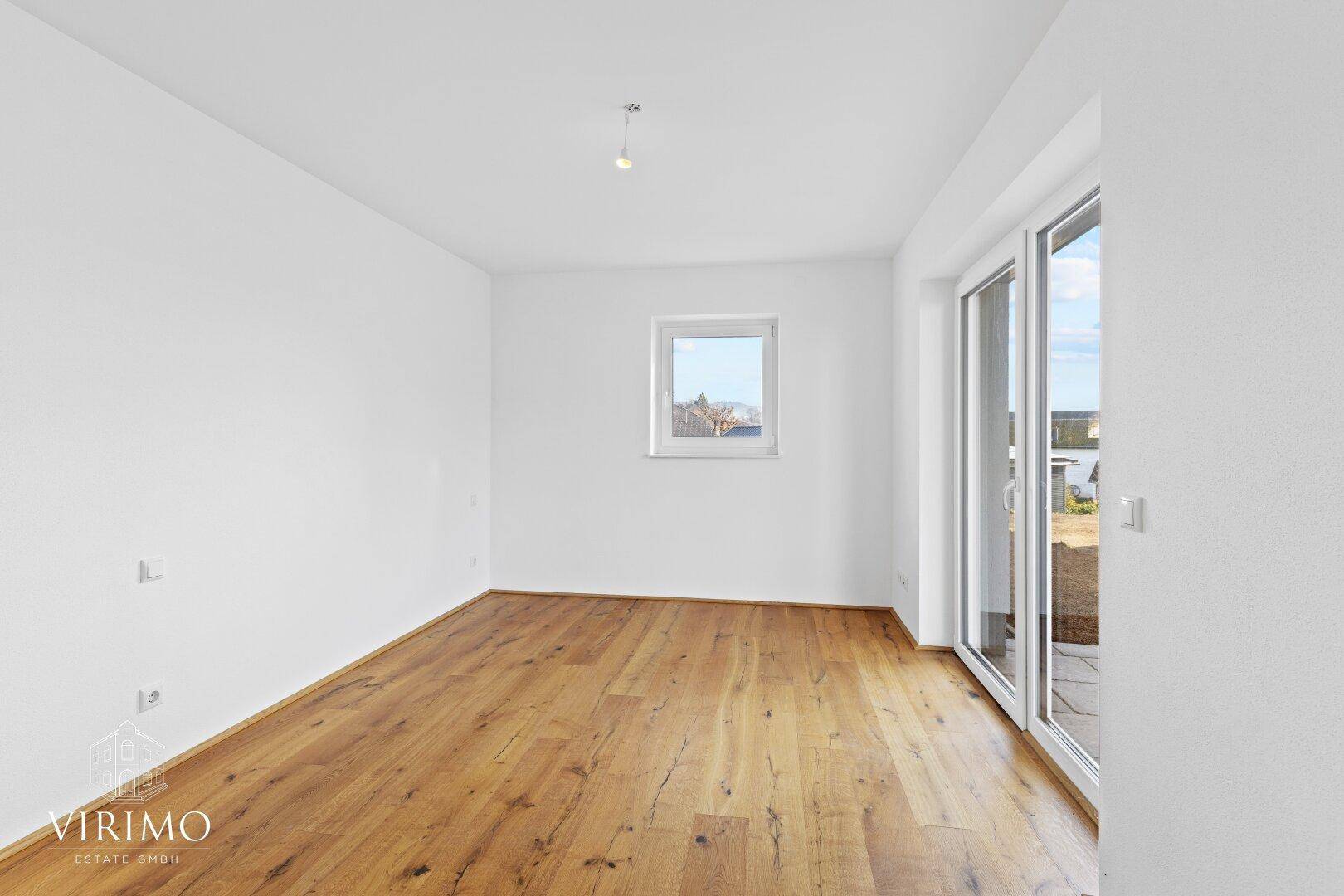 Helles Schlafzimmer mit Holzboden und großem Fenster, das viel Tageslicht hereinlässt.