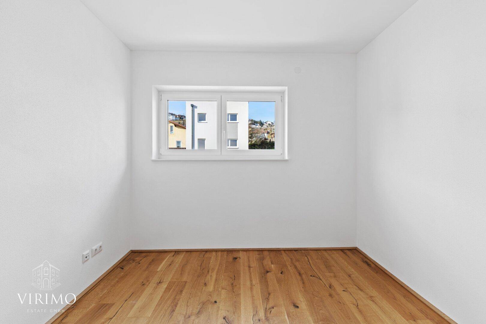 Leeres Zimmer mit Holzboden und Fenster, das einen Blick auf die Nachbargebäude bietet.