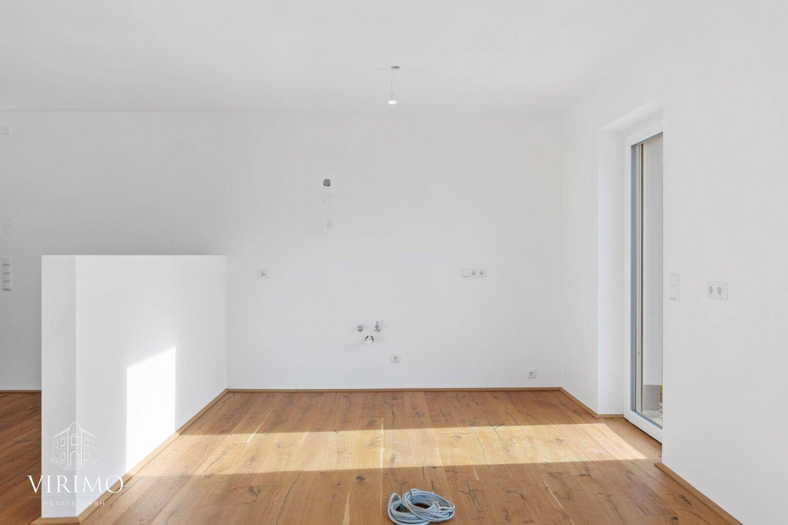 Geräumiger, heller Raum mit Holzboden und vorbereiteten Anschlüssen, ideal für eine offene Küche.