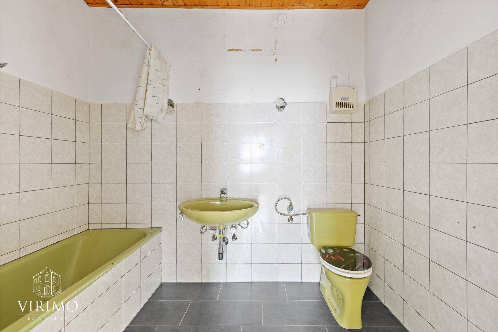 Veraltetes Badezimmer mit grüner Badewanne, Waschbecken und WC, gefliesten Wänden.
