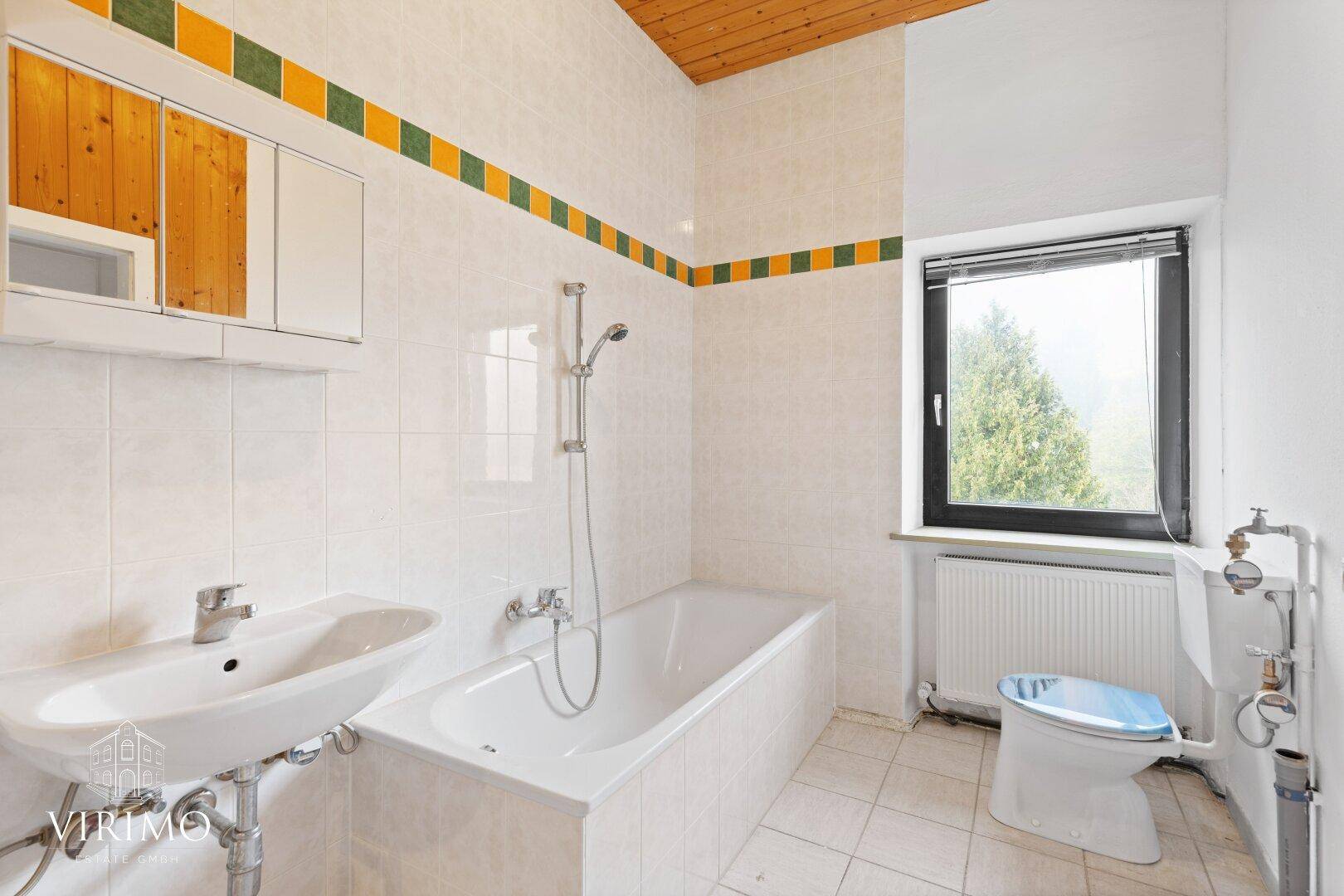 Badezimmer mit Badewanne, Waschbecken, WC und Fenster, teilweise gefliest.