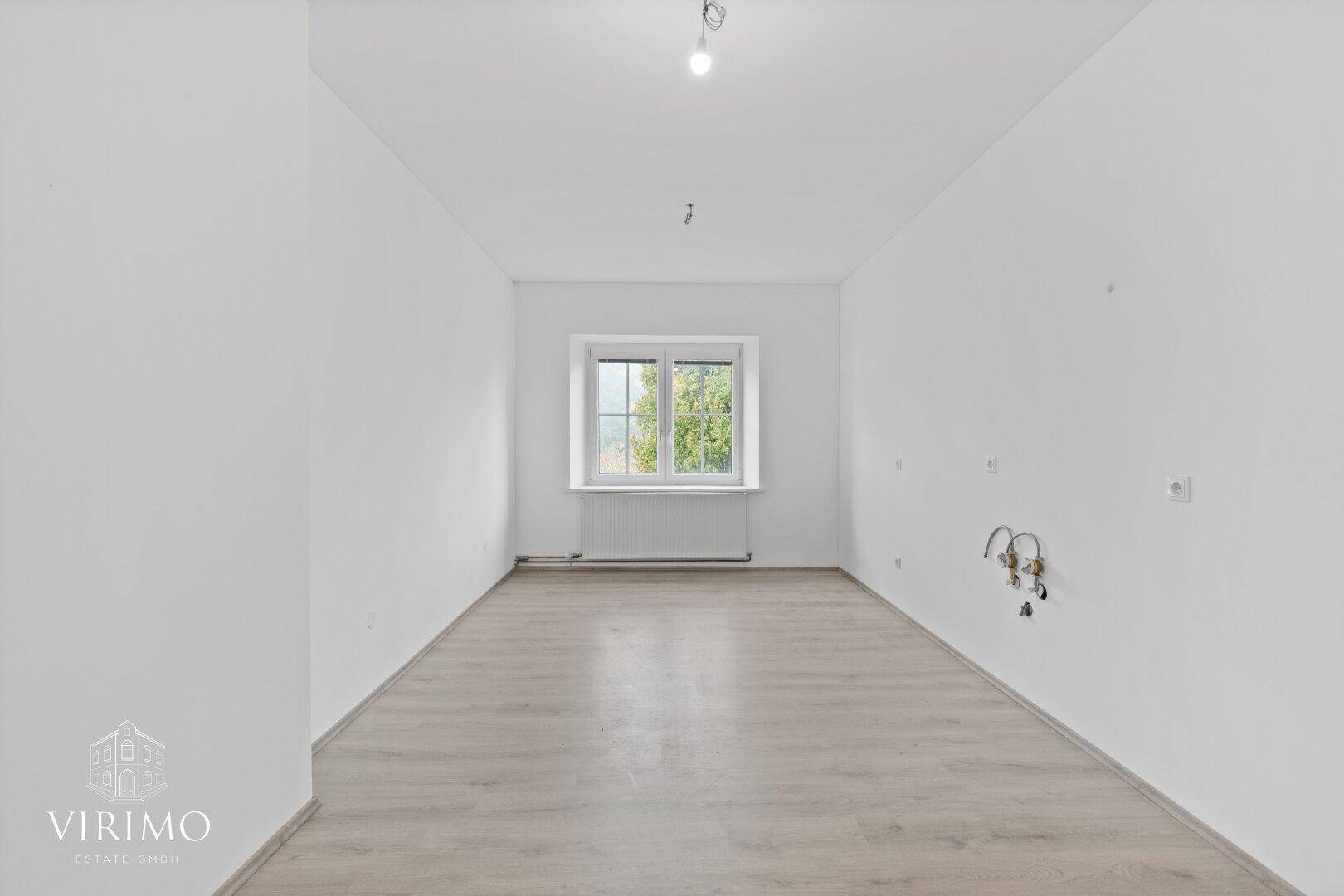 Heller, leerer Raum mit Laminatboden und einem Fenster mit Blick ins Grüne.