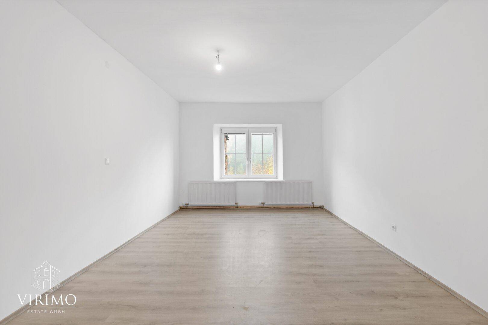 Geräumiger, leerer Raum mit Laminatboden und einem Fenster, das viel Tageslicht hereinlässt.