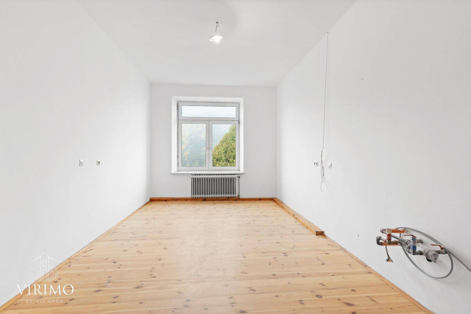 Geräumiger, leerer Raum mit Holzdielenboden und einem Fenster mit Blick ins Grüne.