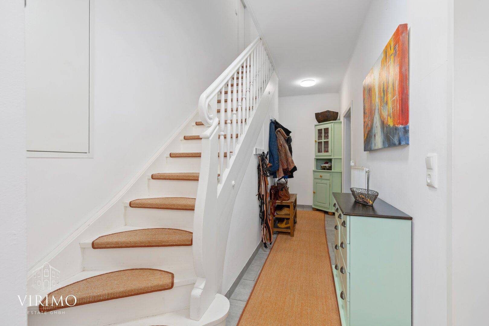 Heller Flur mit weißer Treppe, Garderobe und einem farbenfrohen Kunstwerk an der Wand.