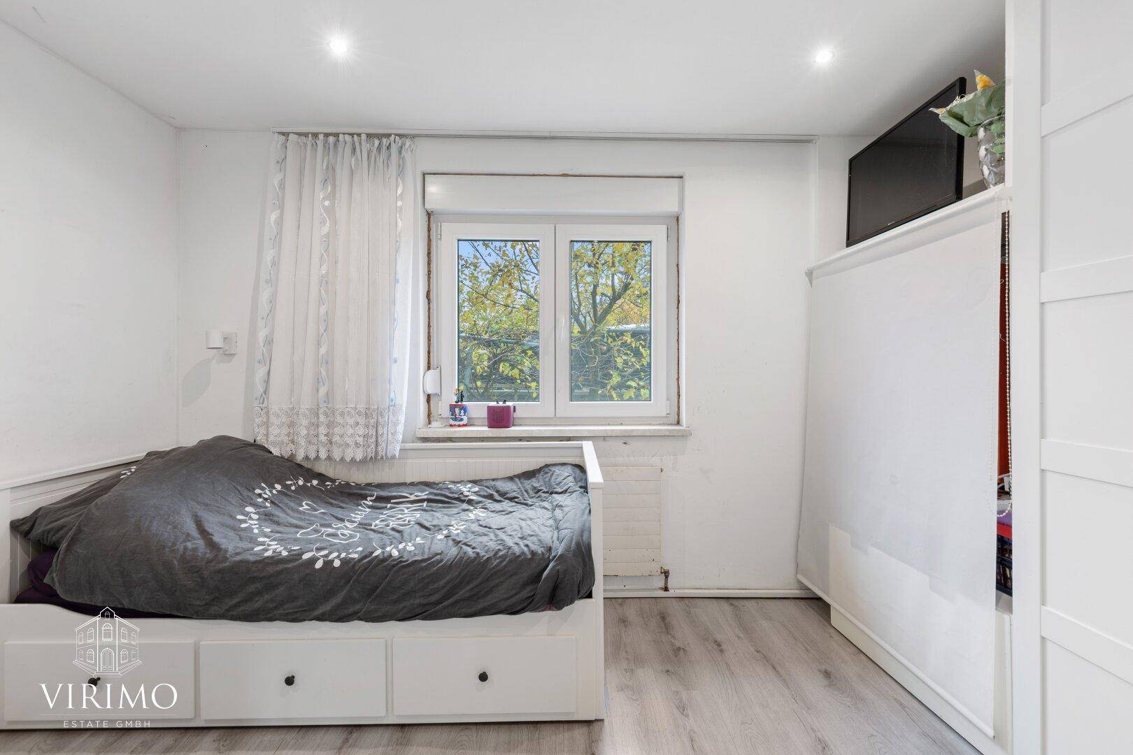Helles Schlafzimmer mit Einzelbett, Schrank und Fenster mit Blick ins Grüne.