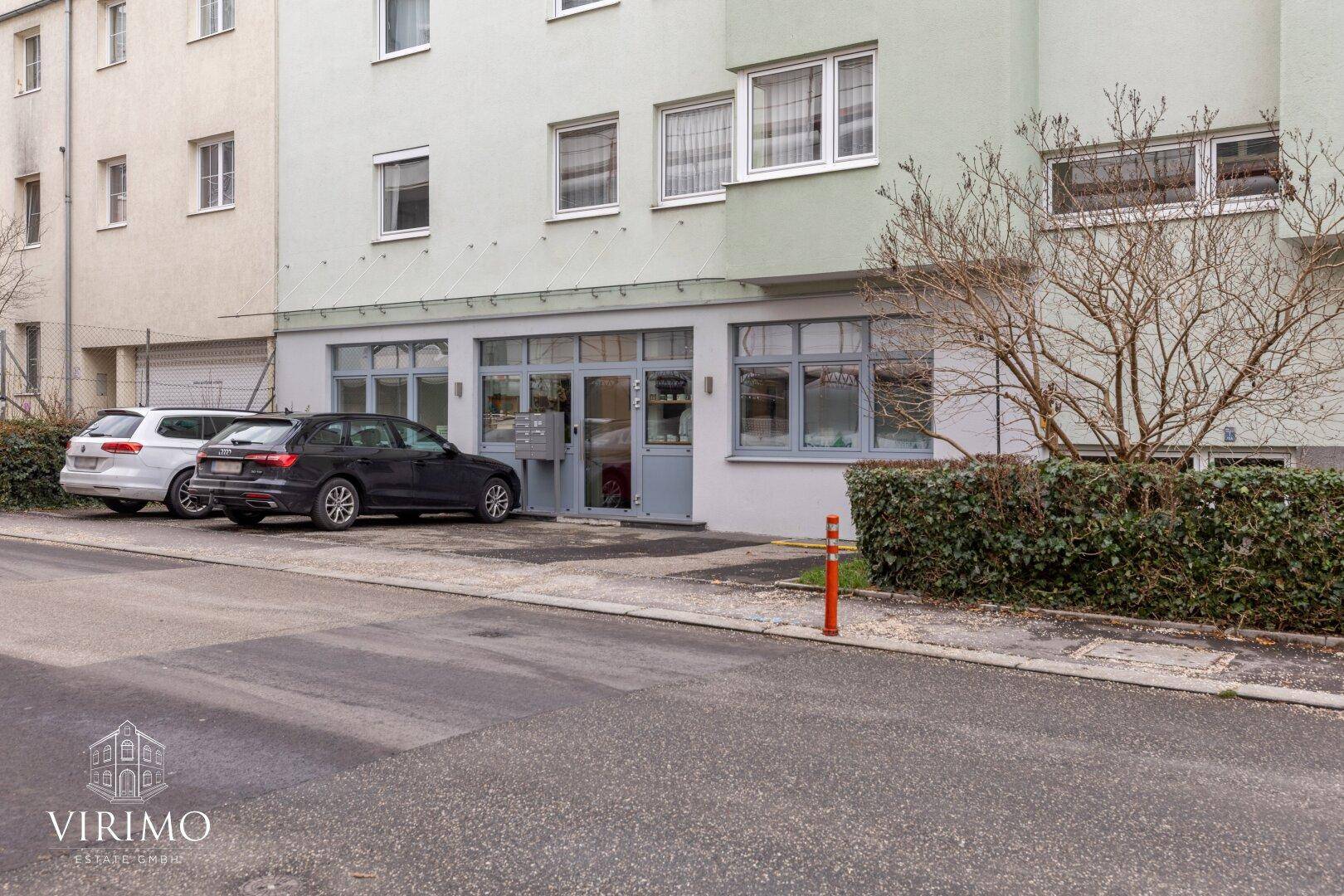 Ansicht des Gebäudeeingangs mit Parkplätzen und einer gepflegten Hecke.