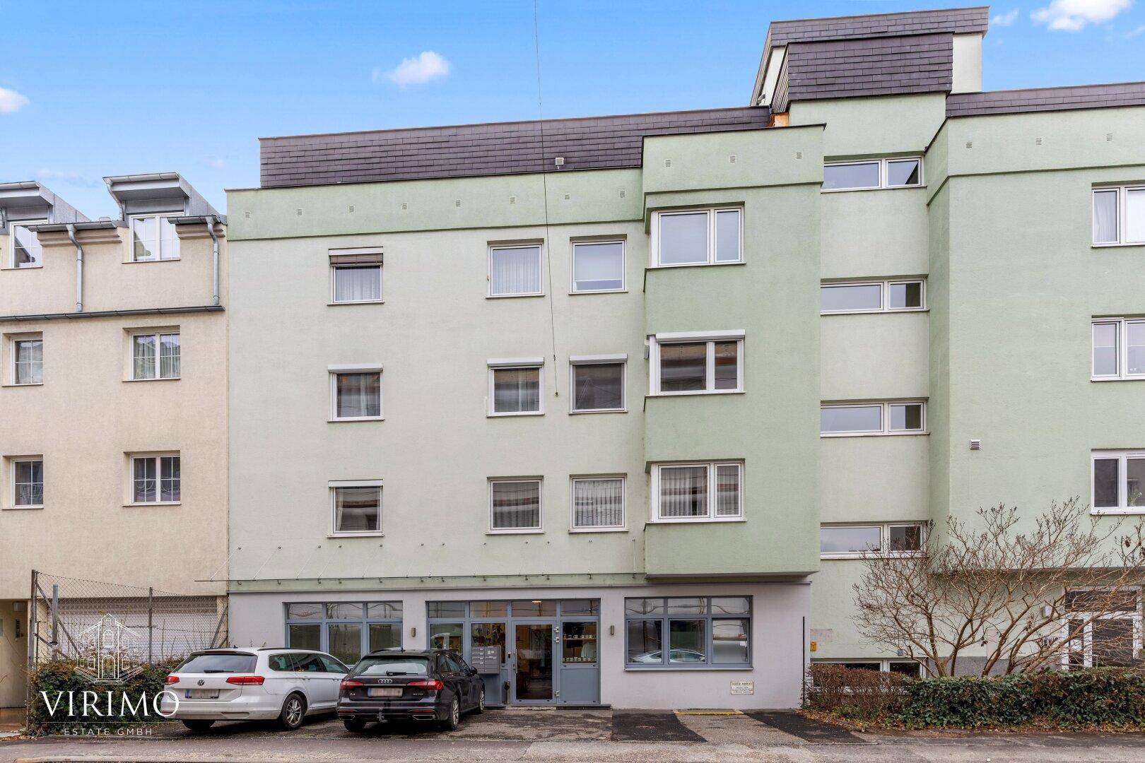 Mehrstöckiges Wohngebäude mit hellgrüner Fassade und Parkplätzen vor dem Eingangsbereich.