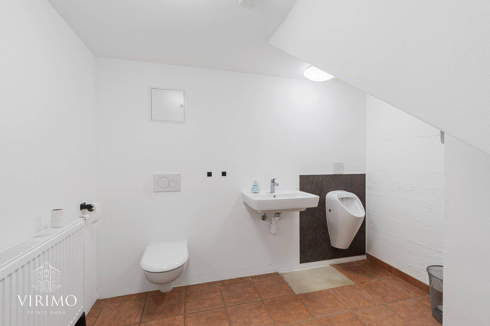 Einfache Toilette mit Urinal, Waschbecken und Fliesenboden im Untergeschoss.