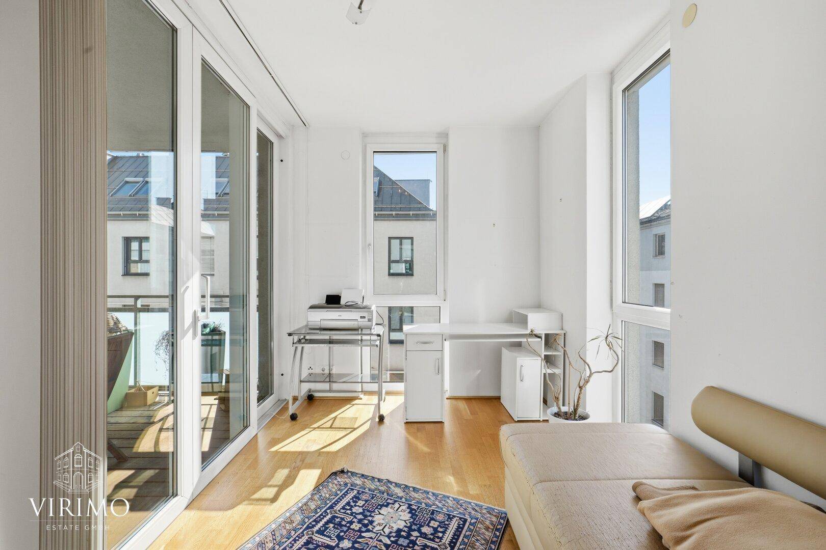 Helles Arbeitszimmer mit Schreibtisch, Sofa und großen Fenstern, eines davon mit Balkonzugang.