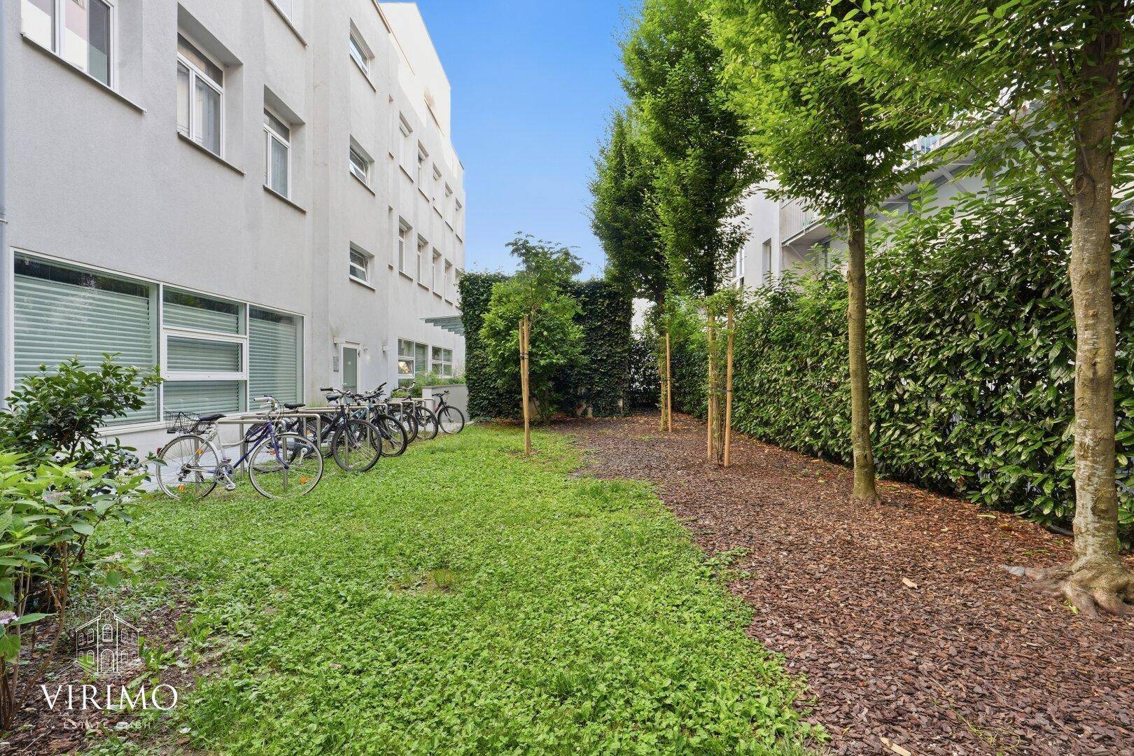 Grüner Außenbereich mit Bäumen, Hecken und Fahrradstellplätzen neben dem Wohngebäude.