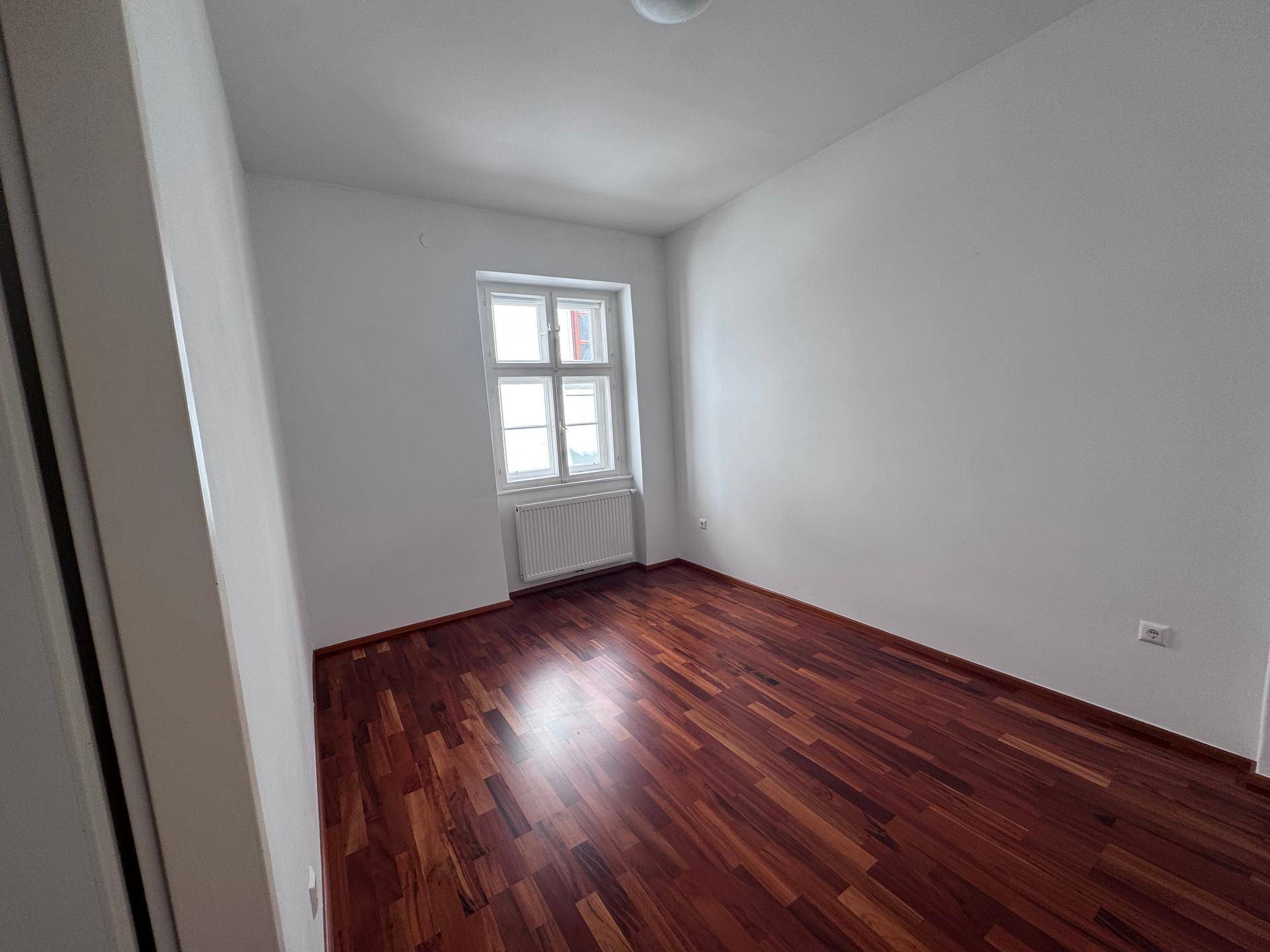 Helles Zimmer mit dunklem Holzboden und einem Fenster, das für natürliche Belichtung sorgt.