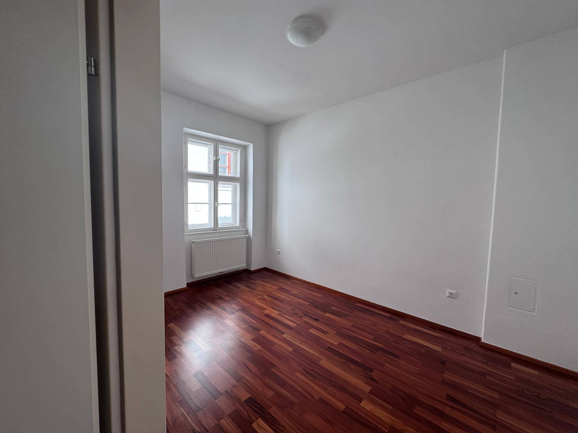 Leeres Schlafzimmer mit Holzboden und einem Fenster, das den Raum mit Licht füllt.