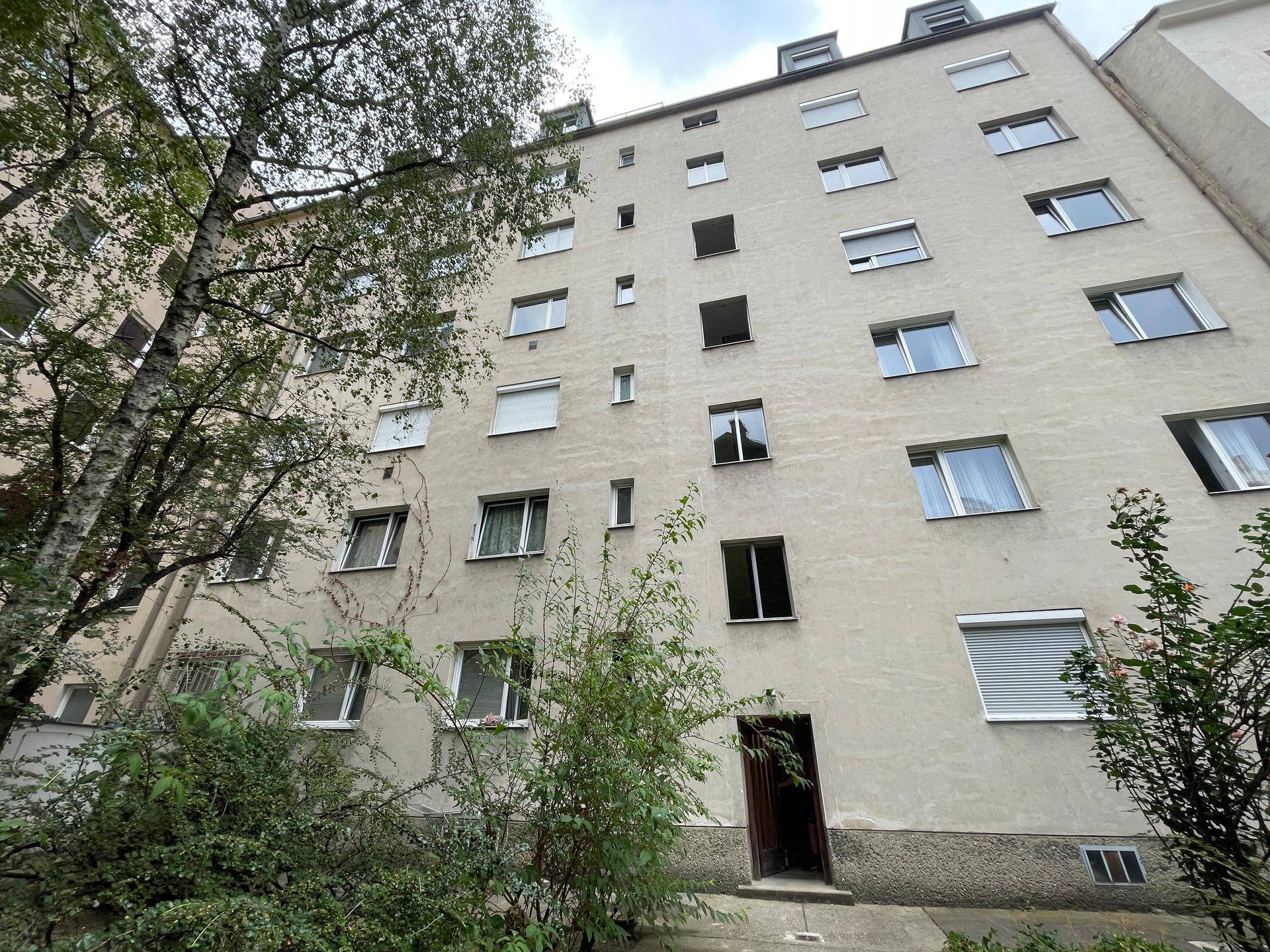Mehrstöckiges Wohngebäude mit zahlreichen Fenstern, Rollläden und Bäumen im Vordergrund.