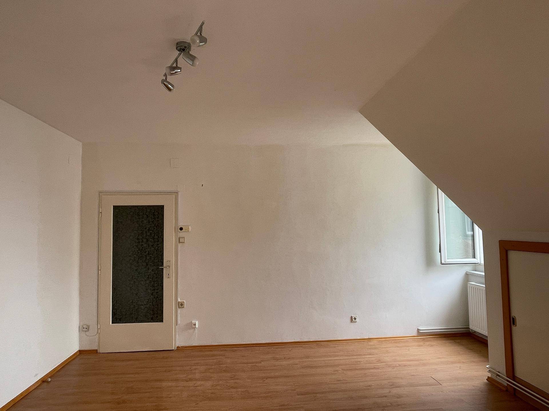 Heller Raum mit Holzboden, schrägen Wänden und einer Tür, die zu weiteren Bereichen führt.