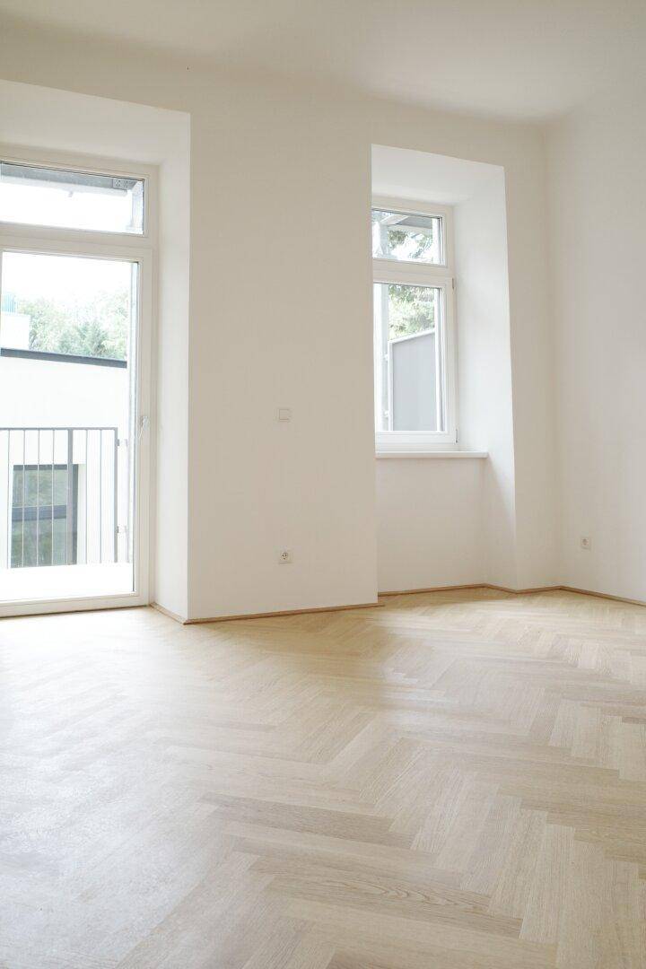Helles, leeres Zimmer mit Fischgrätparkett und Zugang zu einem Balkon.