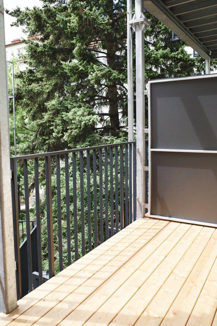 Balkon mit Holzplankenboden und Blick auf grüne Bäume und die Umgebung.