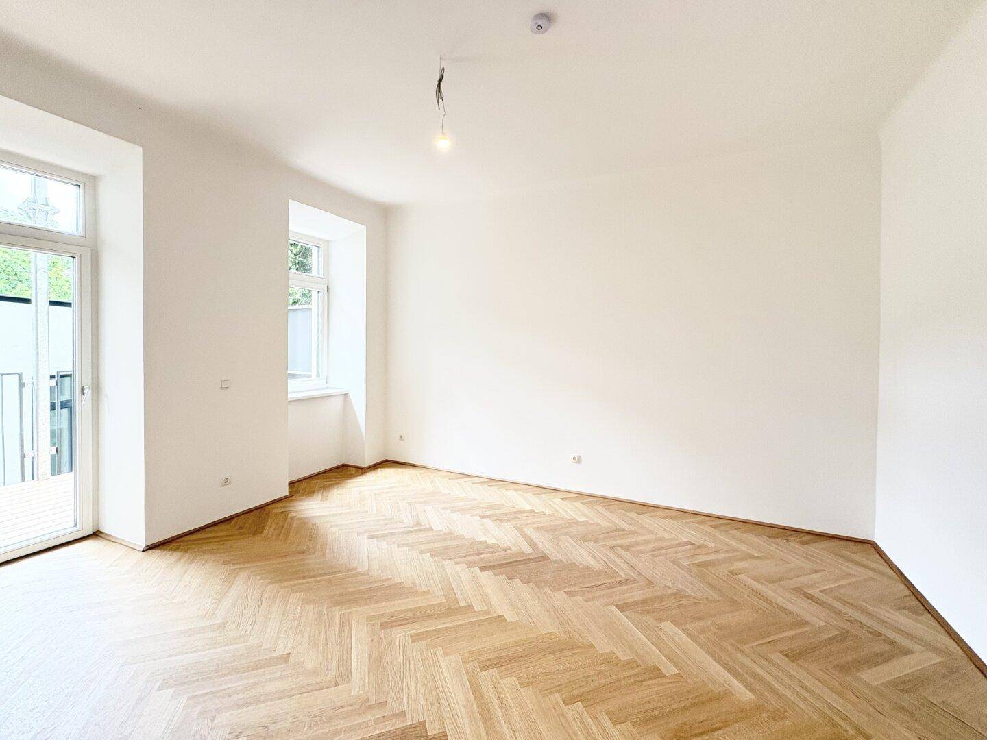 Geräumiges, leeres Zimmer mit Fischgrätparkett und Zugang zu einem Außenbereich.