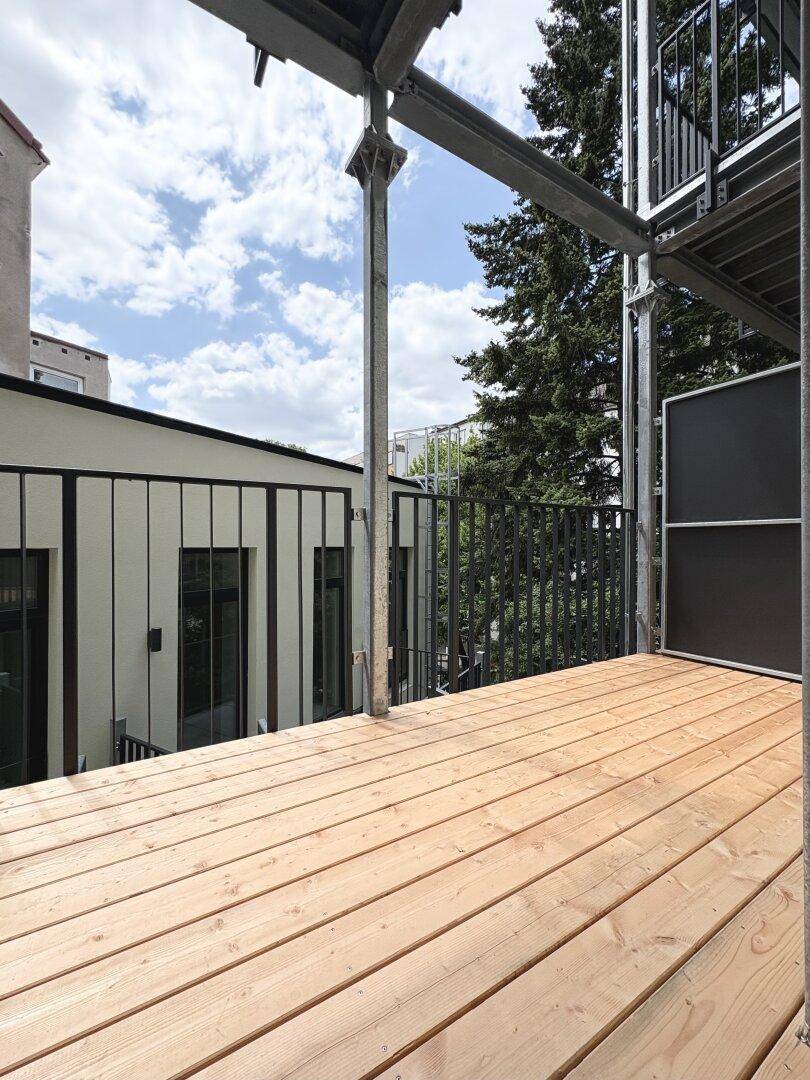 Heller Balkon mit Holzplankenboden und Metallgeländer, umgeben von Bäumen.